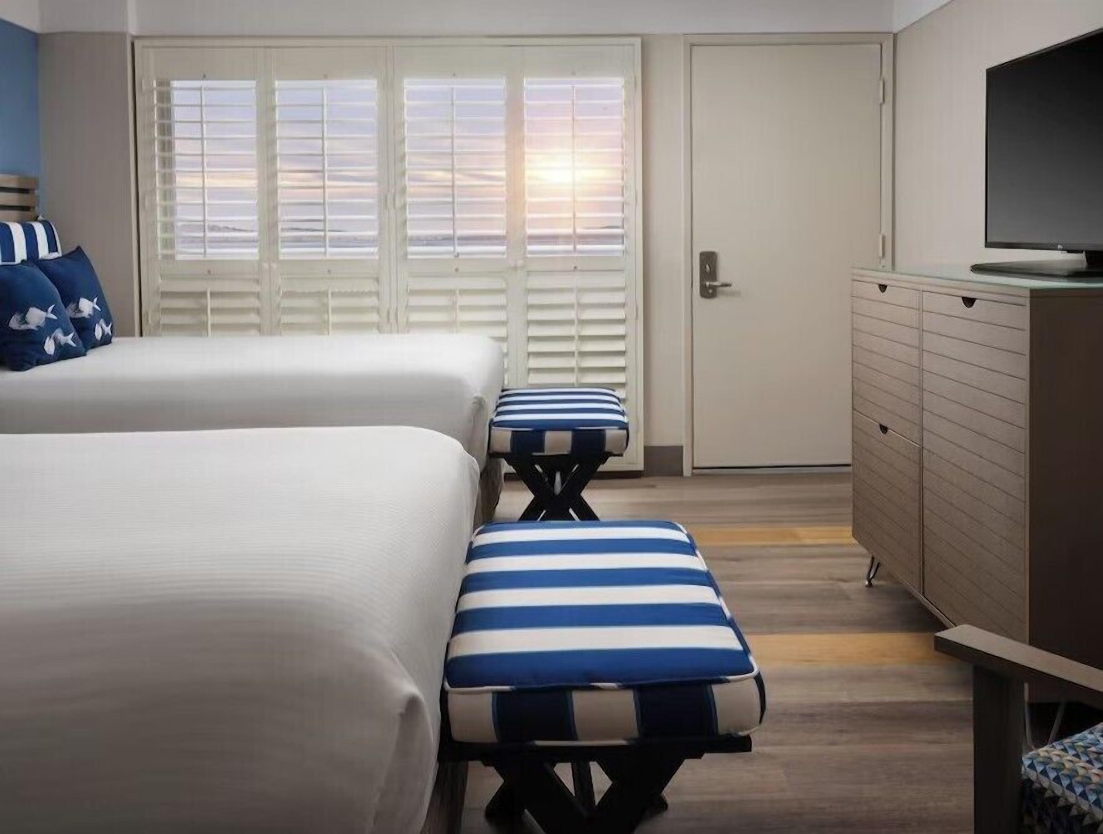 A hotel room with two beds, a bench, a television, and a door with shutters.