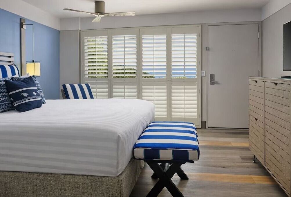 Une chambre à coucher comprenant un lit, un banc, une commode et une porte avec des stores.