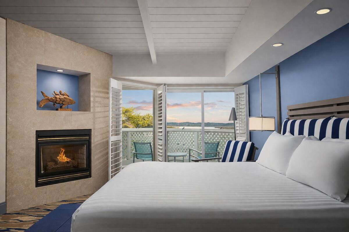A hotel room with a bed, a fireplace, a balcony with chairs, and a view of the landscape.