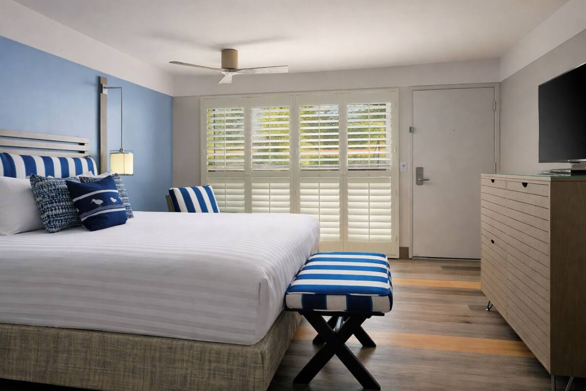 A bedroom with a bed, a bench, a dresser, a ceiling fan, and a window with blinds.