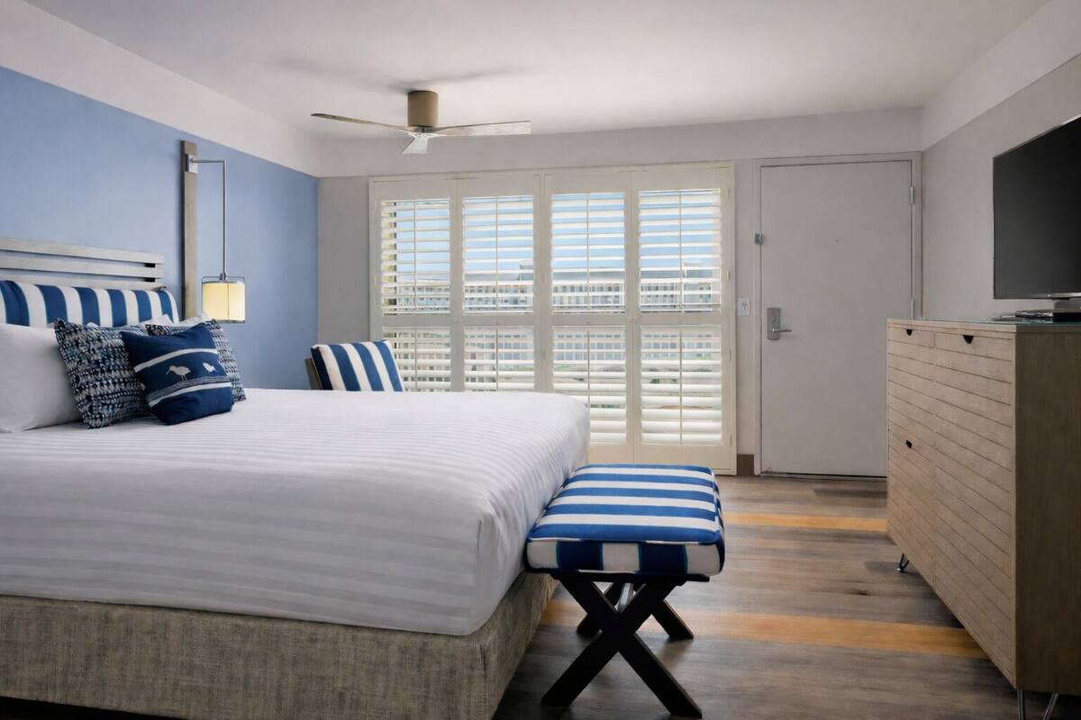 A bedroom with a bed, a bench, a dresser, a television, and a window with blinds.