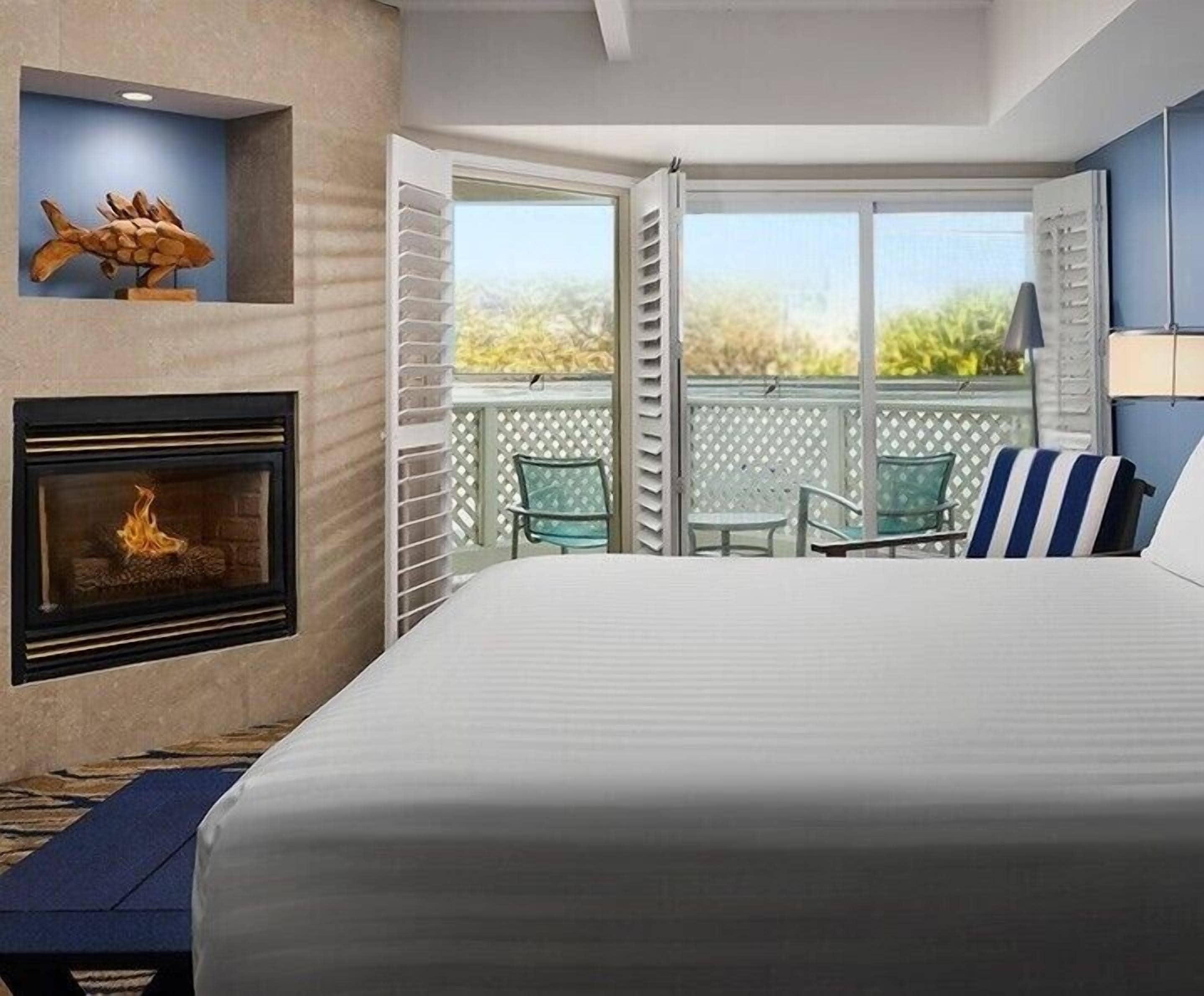 Una habitación de hotel con cama, chimenea, balcón con vistas y una pequeña mesa con sillas.
