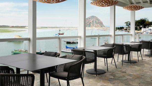 A seaside restaurant with outdoor seating, large windows, and hanging wicker lamps.