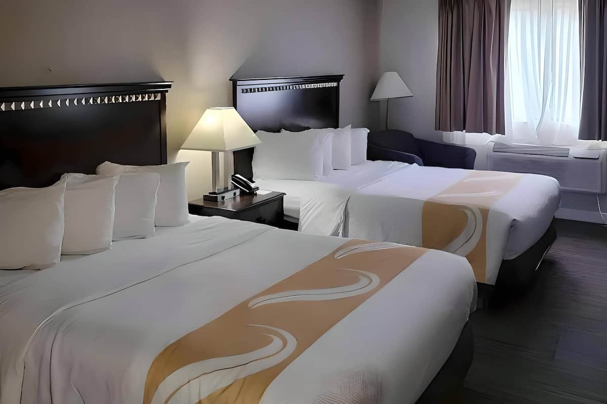 Ein Hotelzimmer mit zwei Betten, einem Nachttisch mit Lampe, einem Schreibtisch mit Stuhl und einem Fenster mit Vorhängen.