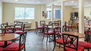 A dining area with multiple tables and chairs, a window with blinds, and a kitchen area in the background.