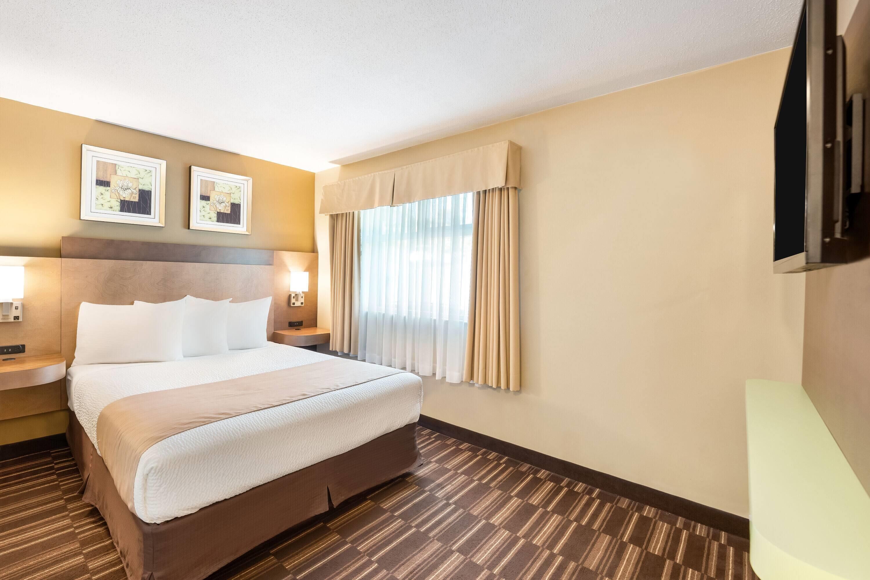 A hotel room with a large bed, two bedside tables with lamps, a flat-screen TV mounted on the wall, and a window with curtains.