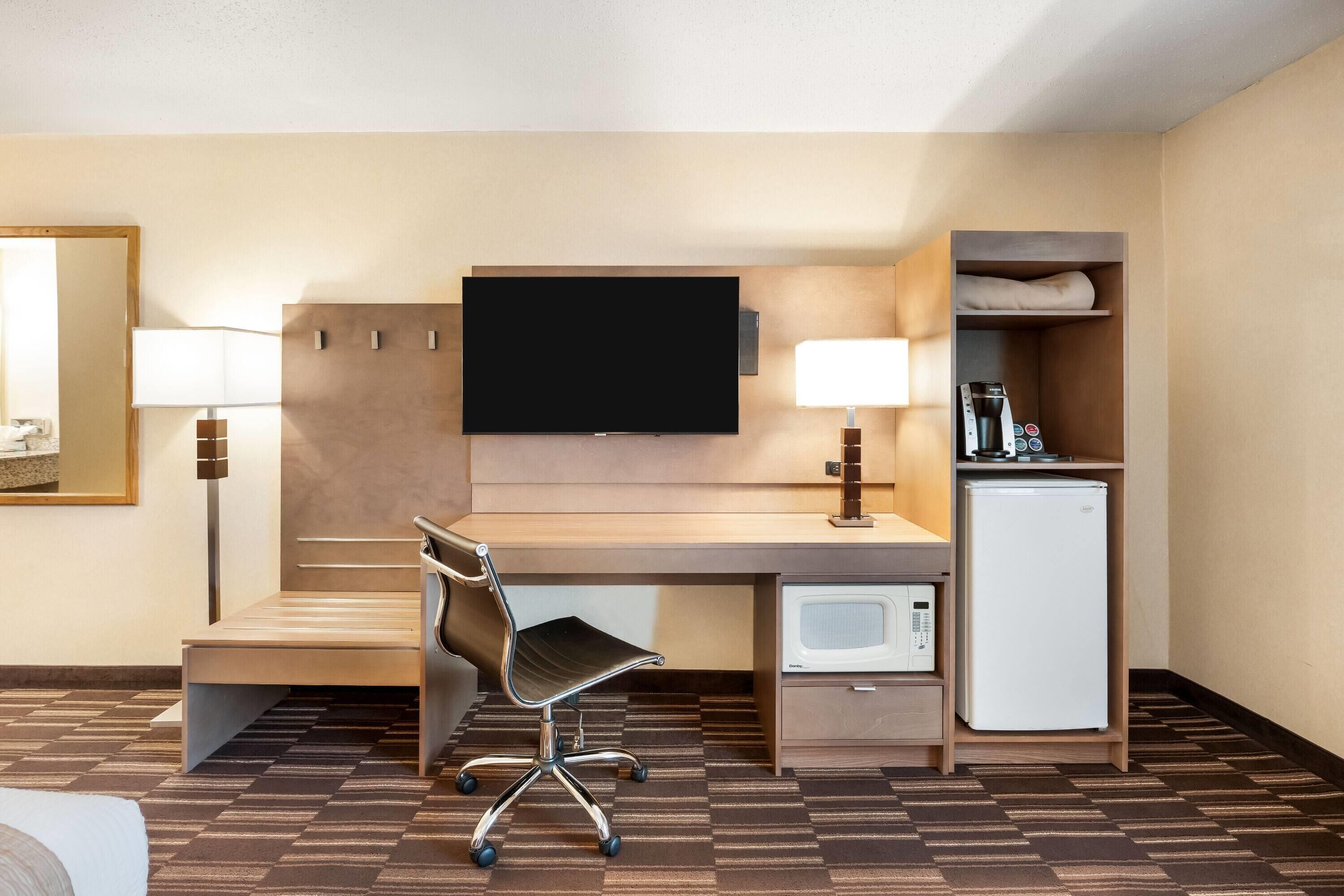 A hotel room with a desk, chair, television, lamp, mini-fridge, and a microwave.