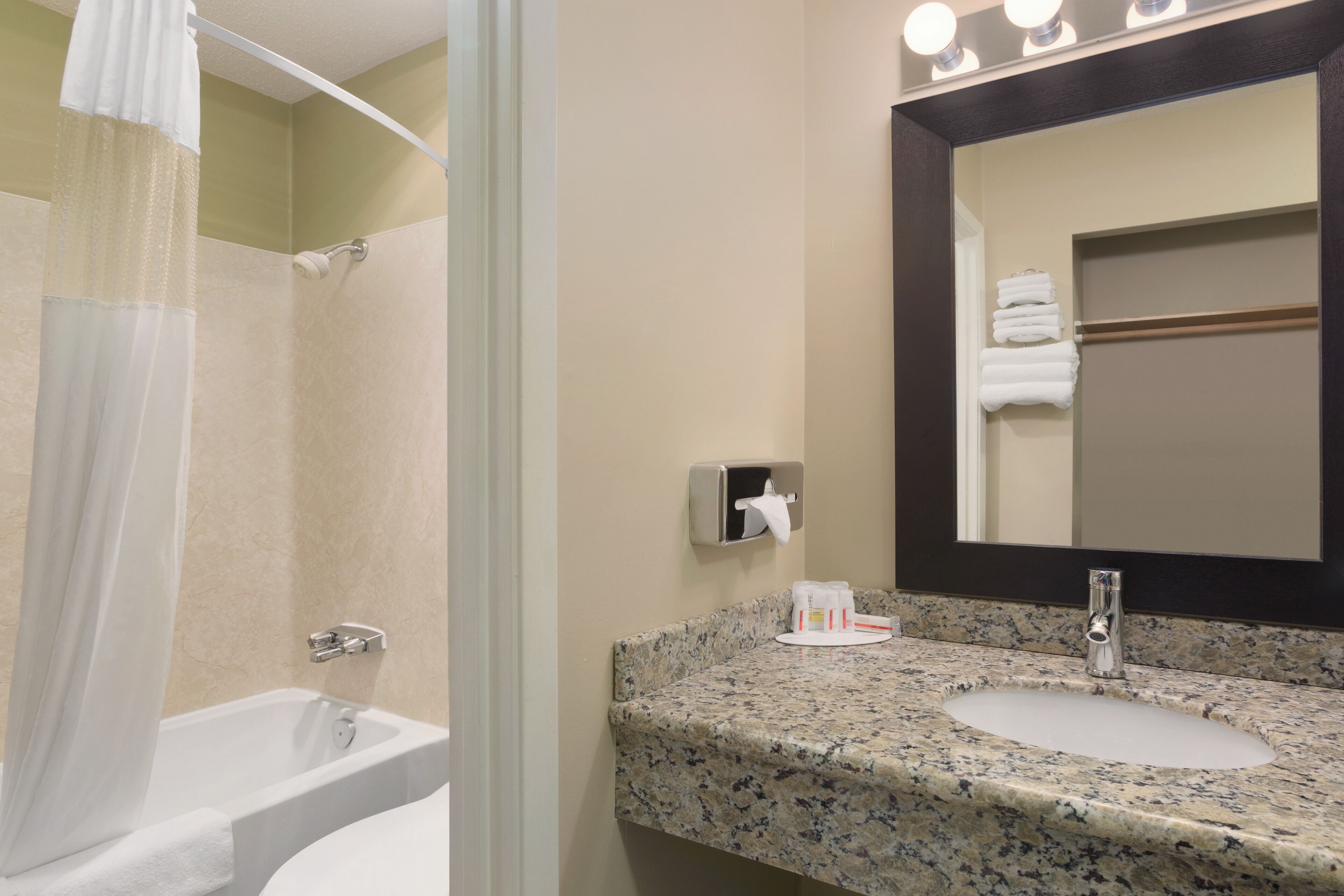 A bathroom with a bathtub, a sink with a granite countertop, a towel dispenser, and a mirror with lights above it.
