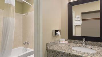 A bathroom with a bathtub, a sink with a granite countertop, a towel dispenser, and a mirror with lights above it.