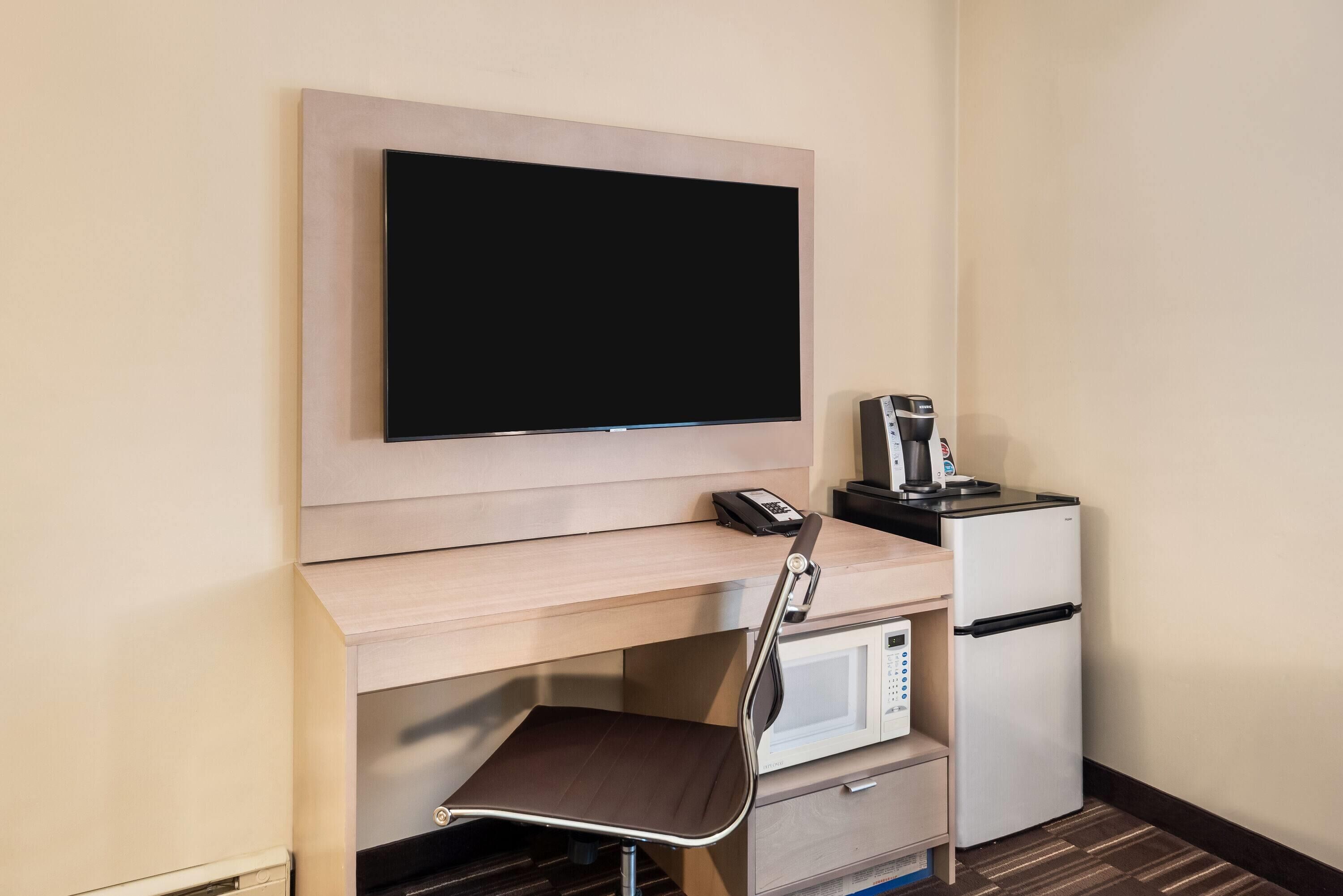 A hotel room with a flat-screen TV mounted on the wall, a microwave, a refrigerator, a chair, and a coffee maker on a small table.