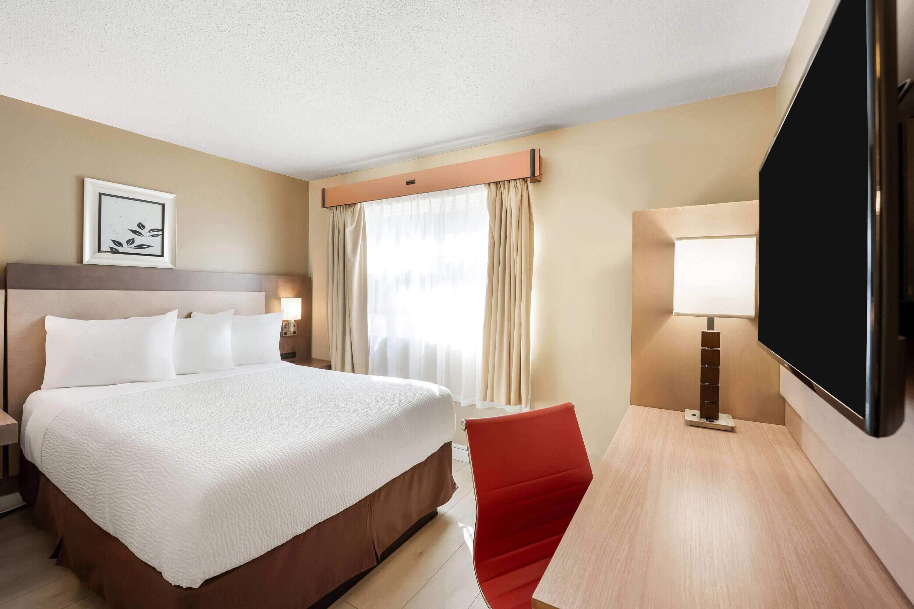 A hotel room with a large bed, a desk with a TV, a red chair, and a window with curtains.