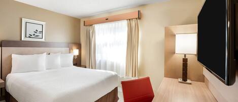 A hotel room with a large bed, a desk with a TV, a red chair, and a window with curtains.