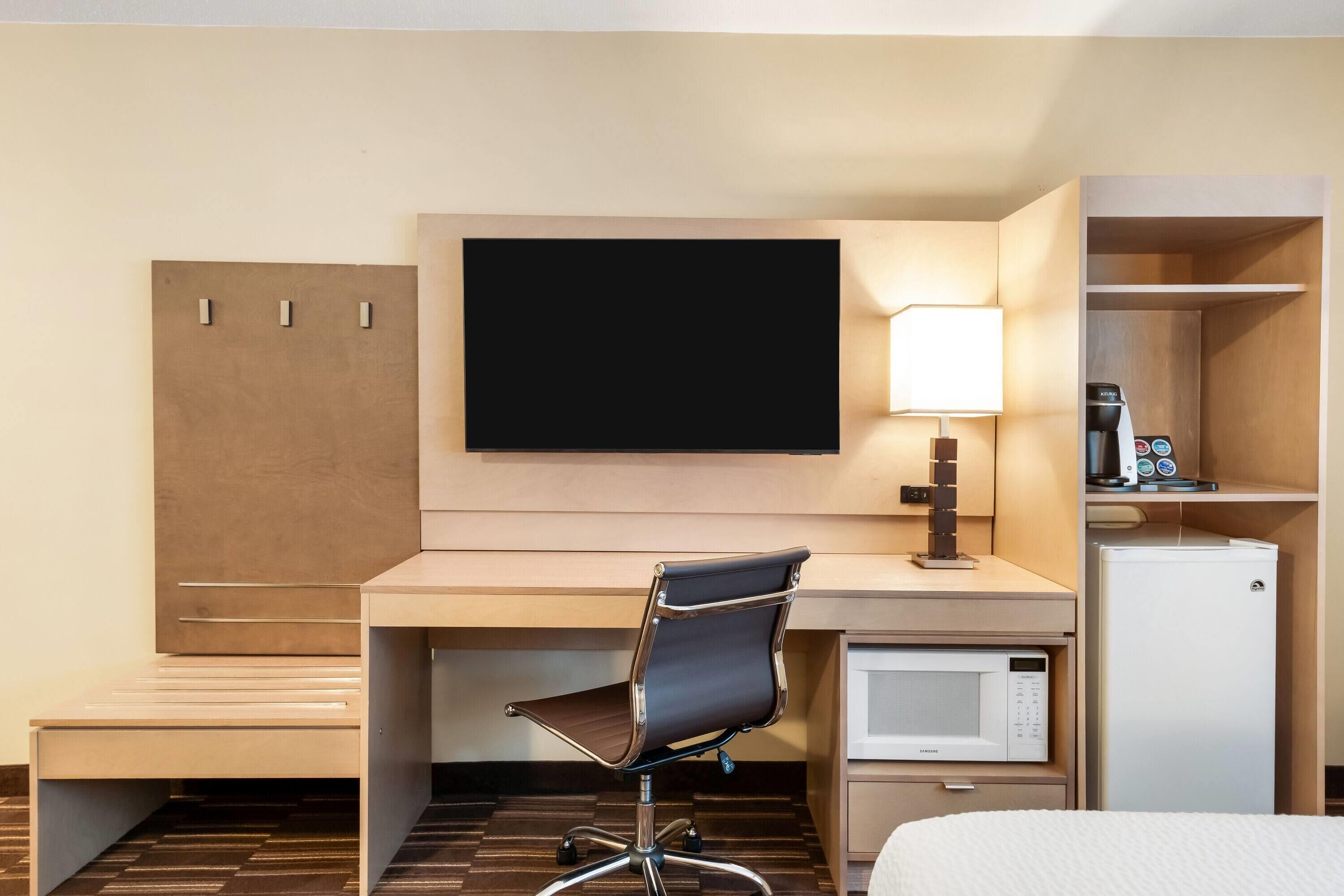 Habitación de hotel con escritorio, silla, televisor, lámpara, minibar y cama.
