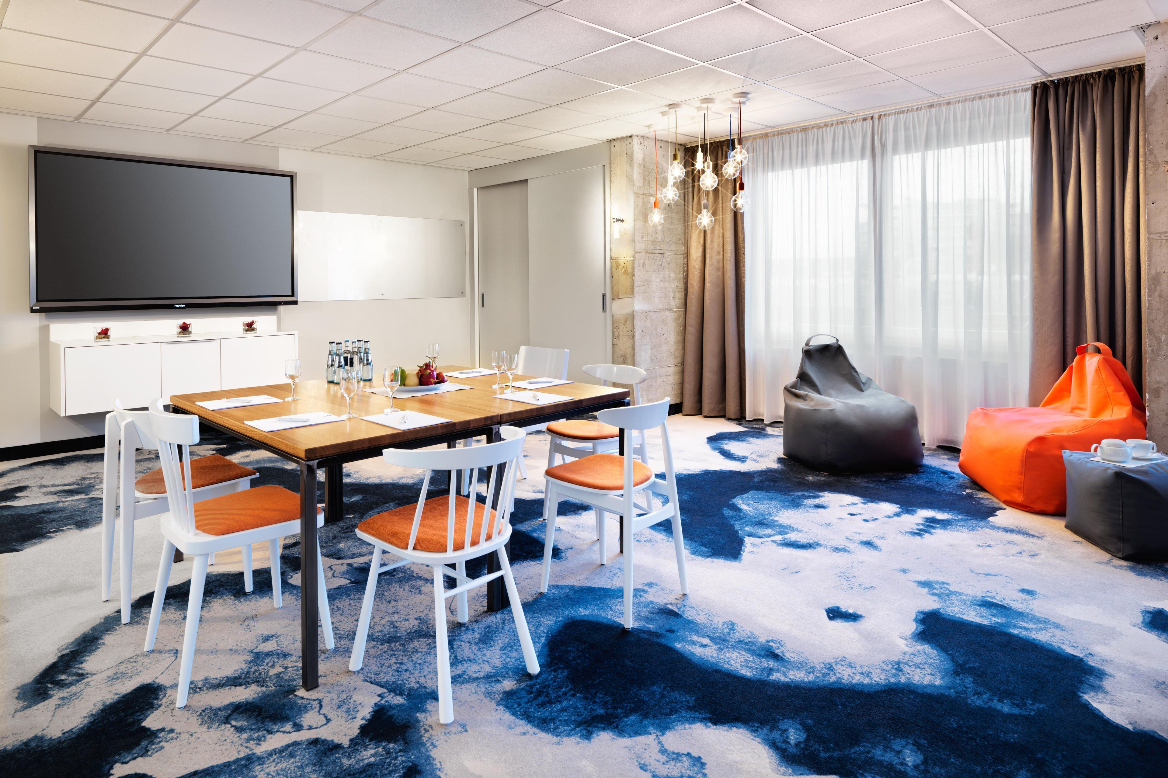 Habitación de hotel moderna con un televisor de pantalla plana grande, una mesa de comedor de madera con sillas blancas, una alfombra estampada en azul y negro, y un área de descanso con puffs tipo frijol naranja y un pouf gris.