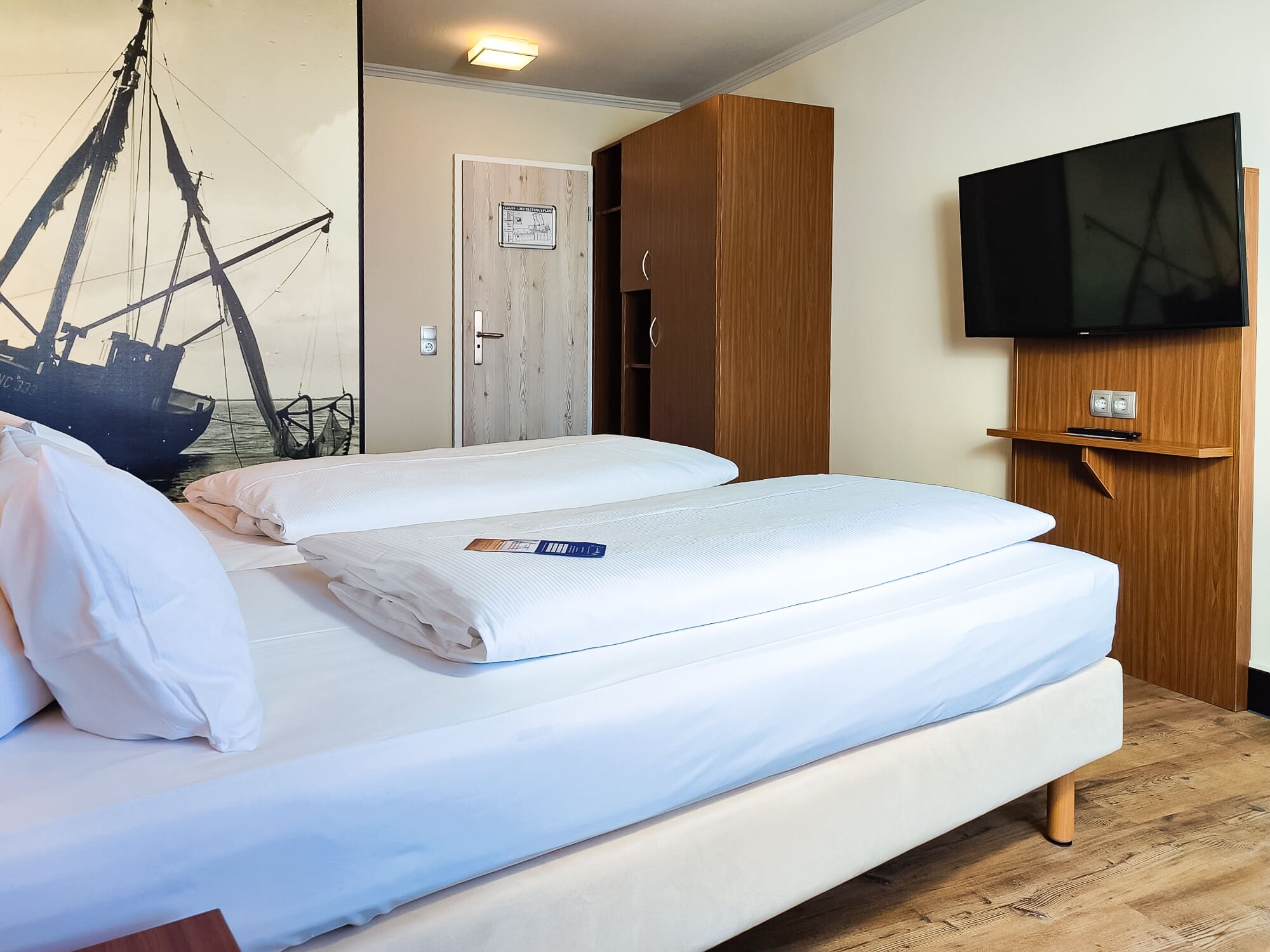 A double bed with white linens, a wooden wardrobe, a television mounted on the wall, and a wooden floor.