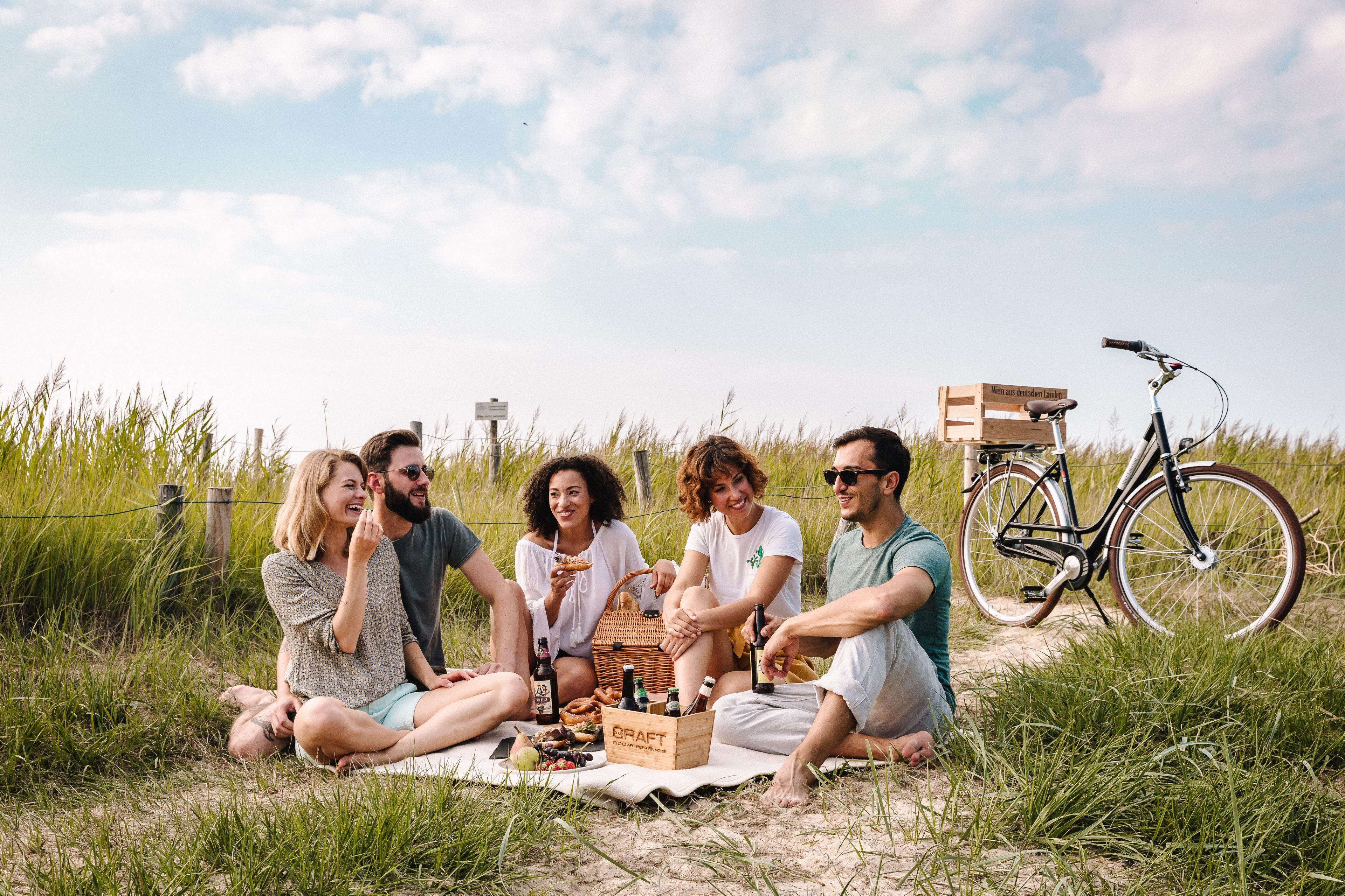 Un grupo de personas sentadas sobre una manta al aire libre, con una bicicleta y un cajón de madera al fondo.