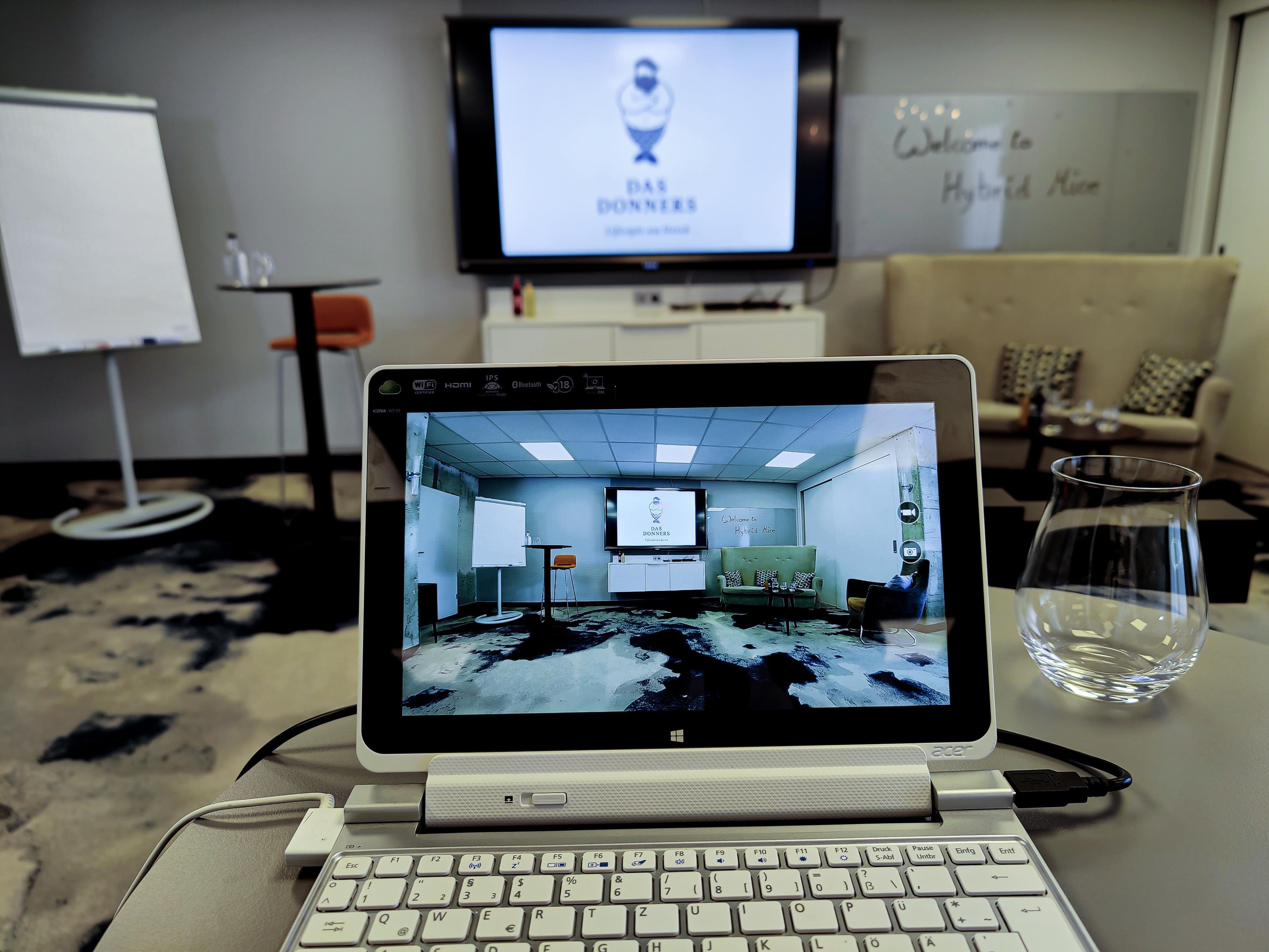 Ein Laptop auf einem Schreibtisch mit Blick auf einen Raum, der eine Couch, eine Whiteboard und einen Fernseher mit einem Logo beinhaltet.
