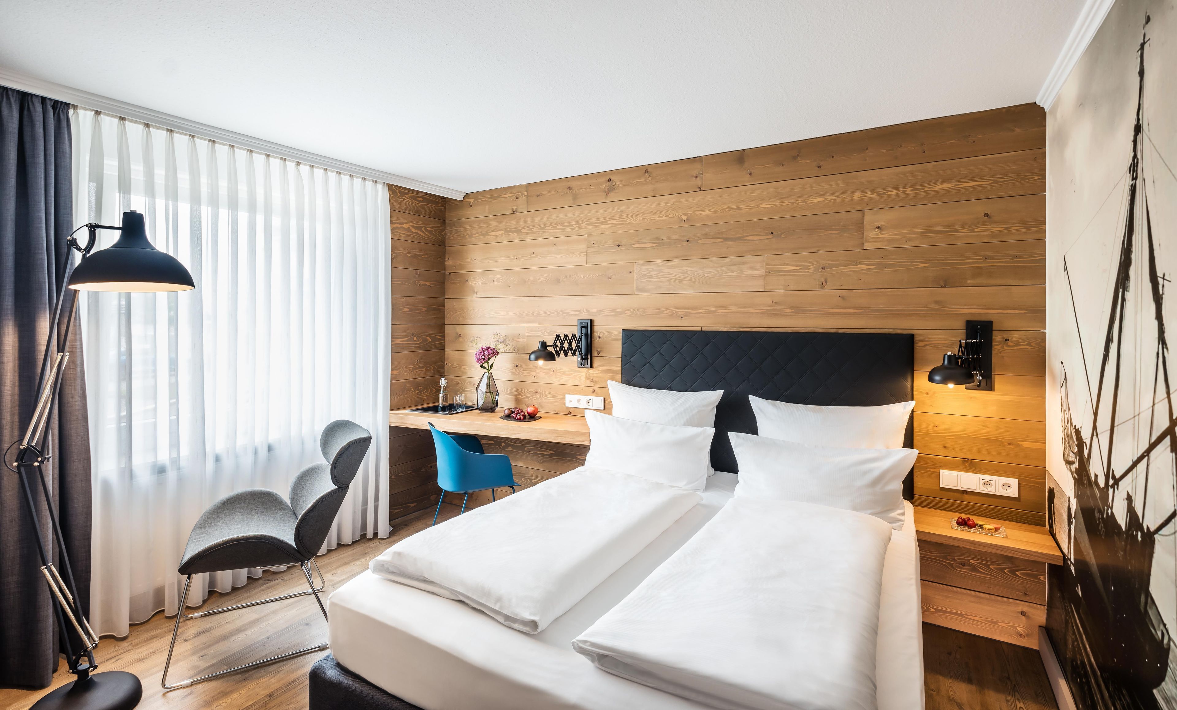 Habitación de hotel con cama, escritorio, silla y un ventanal con cortinas.
