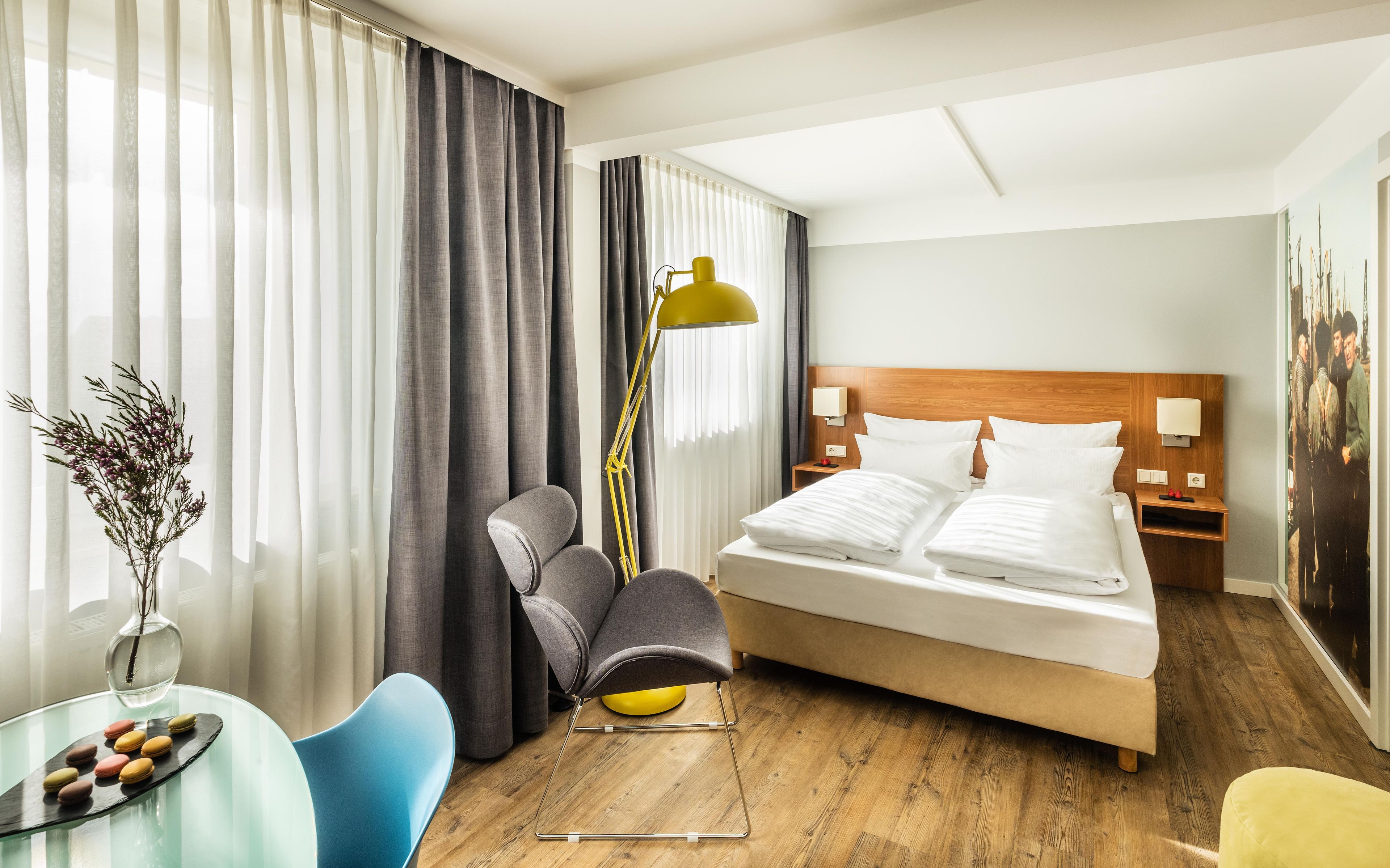 A hotel room with a bed, a desk with a chair, a lamp, and a vase with flowers.