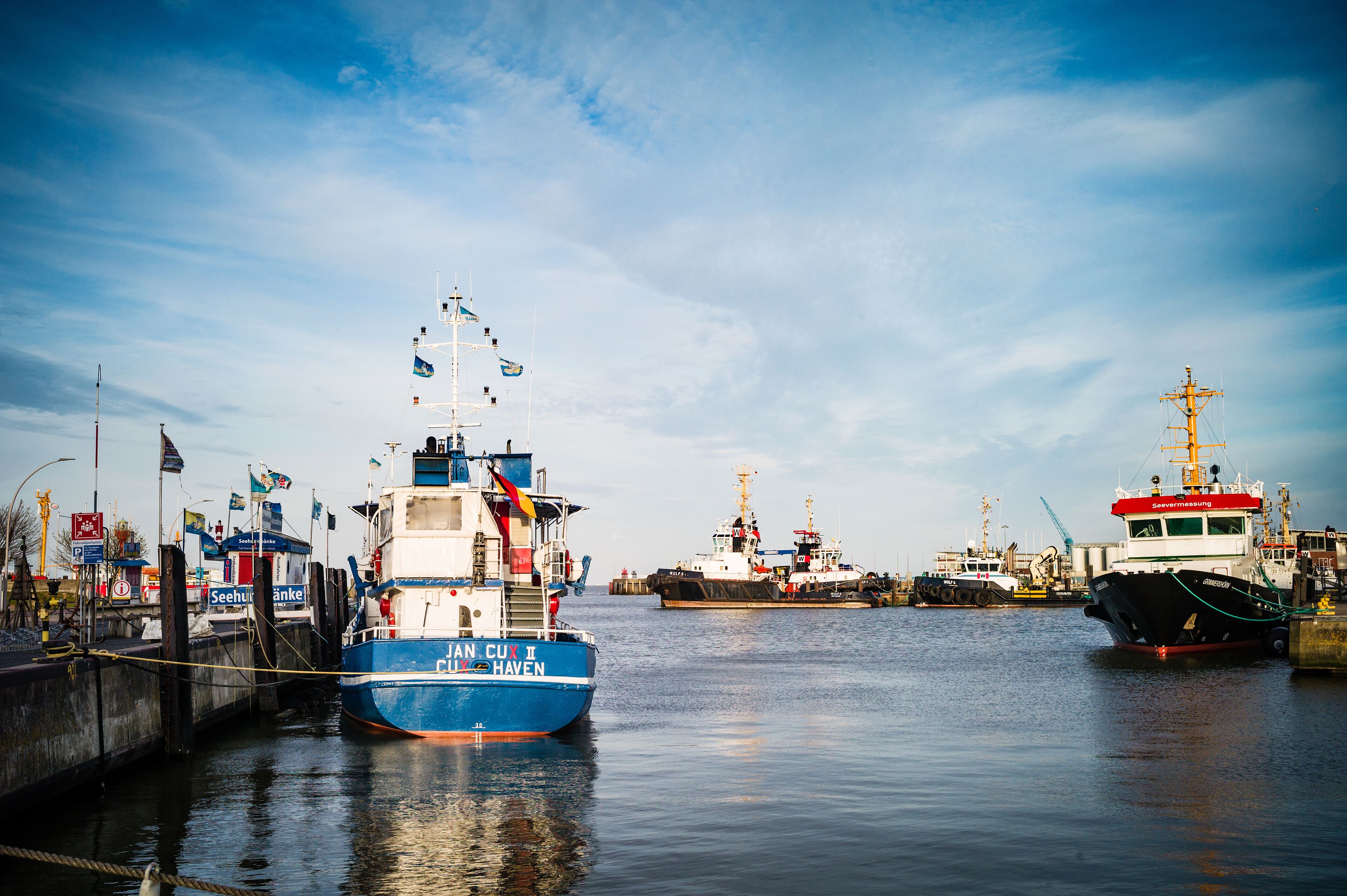 Un puerto con varias embarcaciones amarradas, incluyendo un barco azul llamado "Cuan Cux I" y un barco rojo llamado "Seehafen".