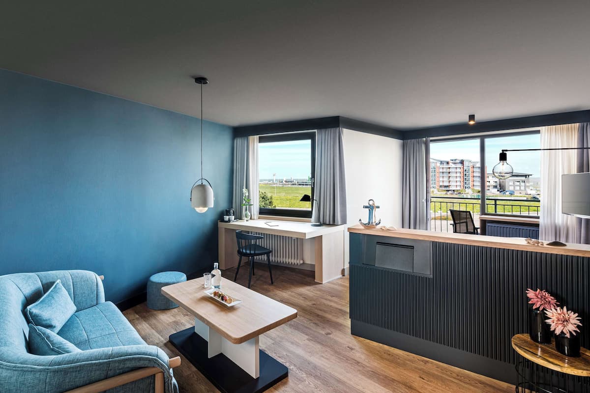 A modern hotel room with a blue wall, a cozy seating area, a small dining table, a balcony with a view of the city, and a large window.