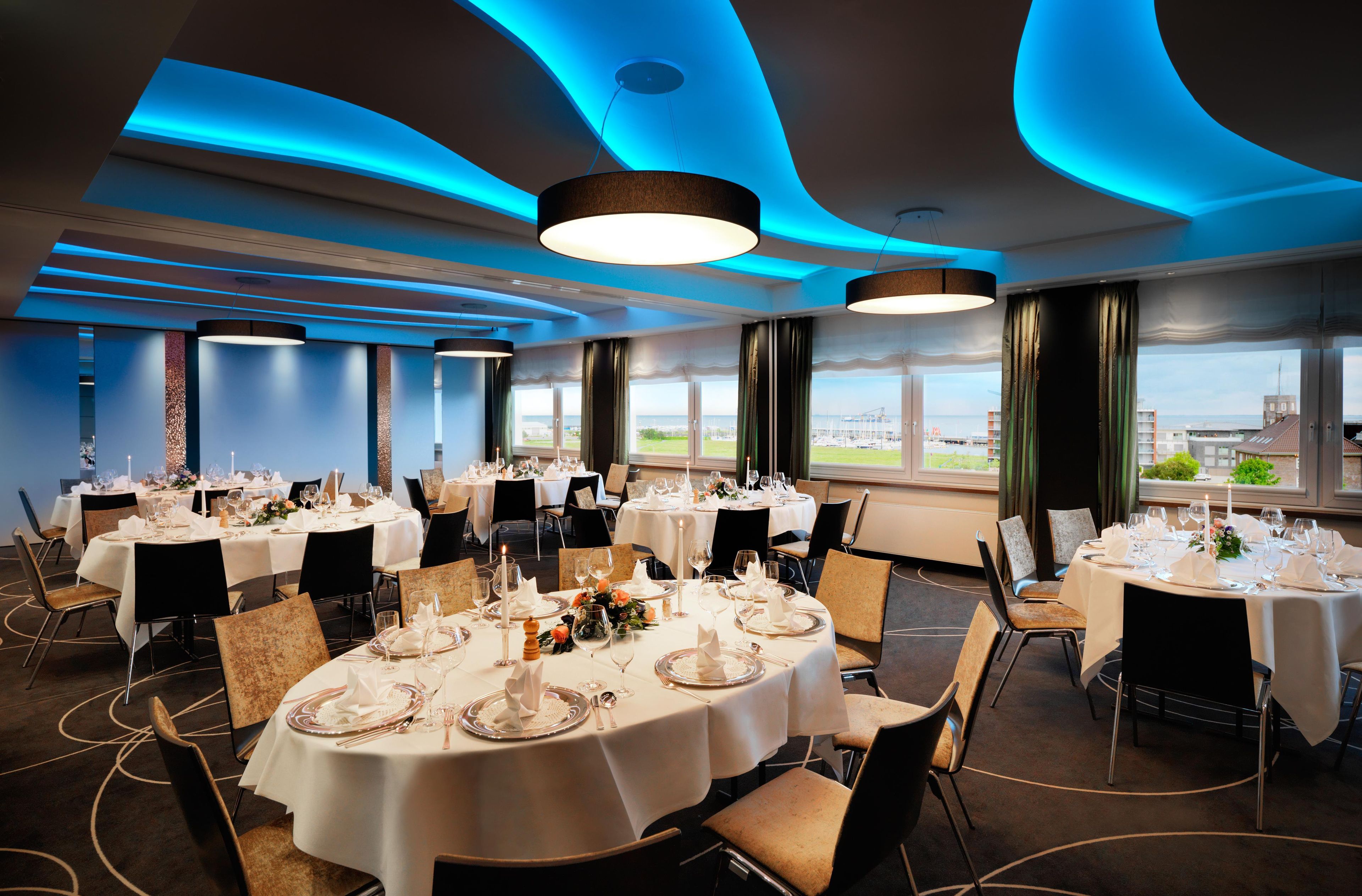 A hotel dining room with round tables set for a meal, large windows with a view, and blue ceiling lighting.