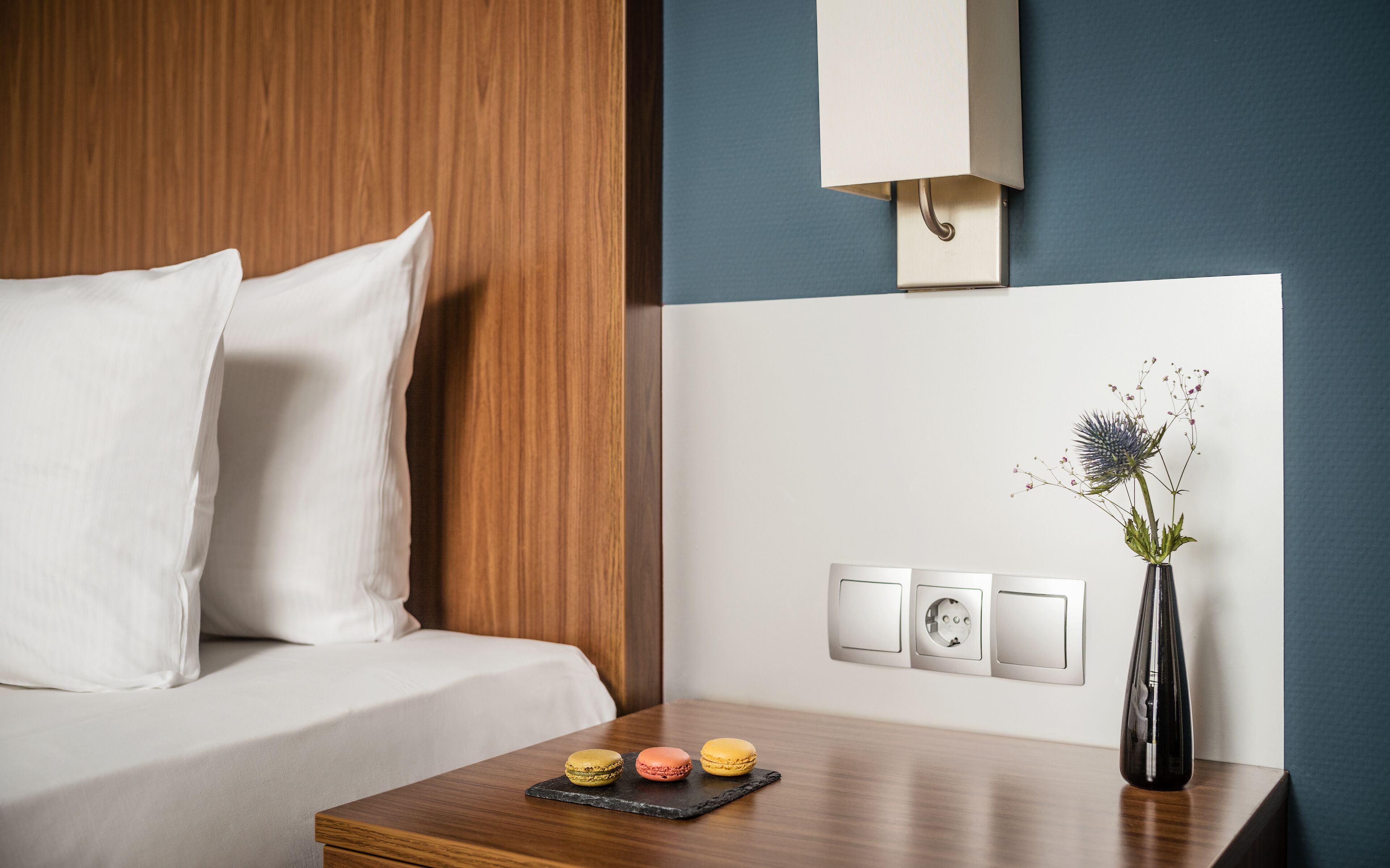 A hotel room with a wooden headboard, white bedding, a bedside table with a vase and flowers, and a wall-mounted light fixture.