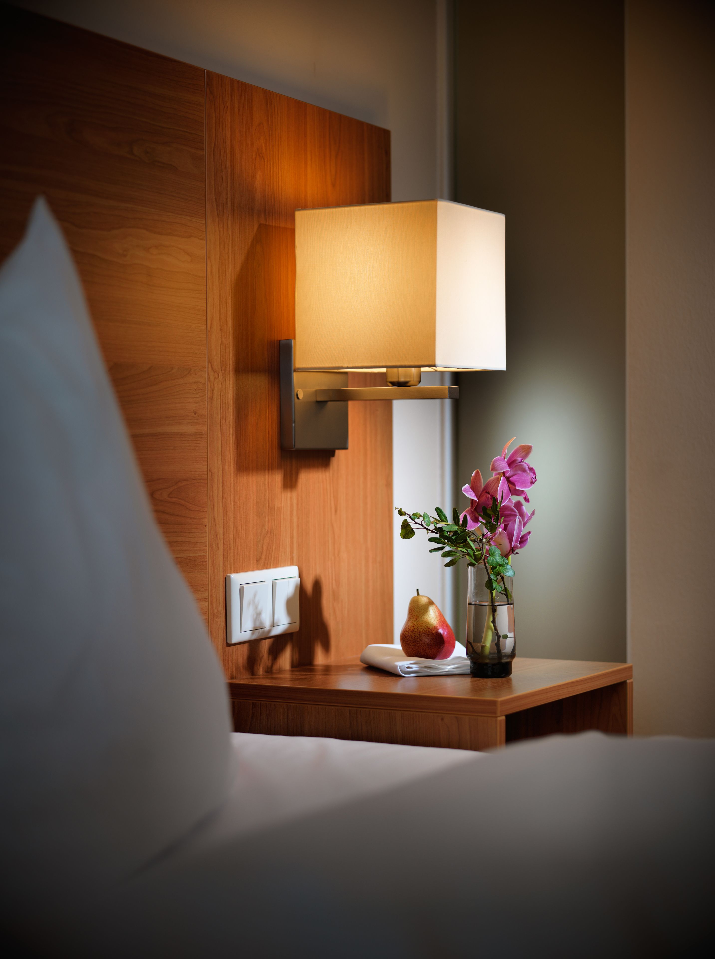 A neatly made bed with a white pillow, a bedside table with a vase of flowers, a pear, and a lamp.