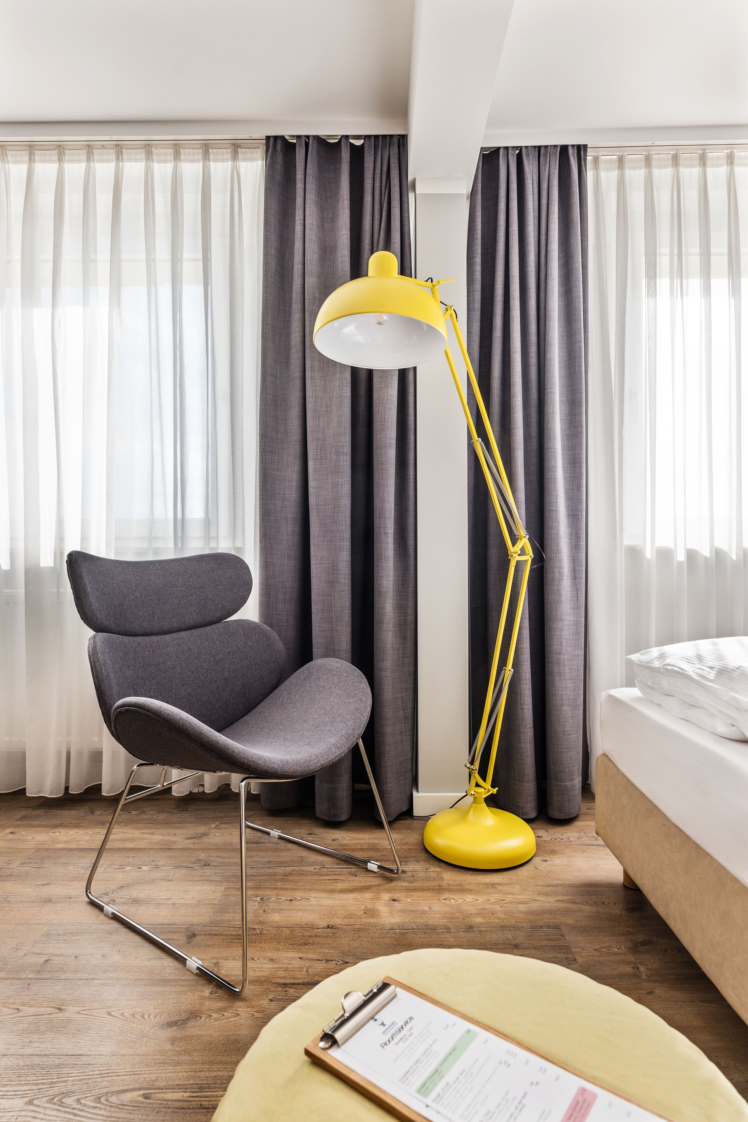 A modern hotel room with a wooden floor, a grey upholstered chair, a yellow floor lamp, and a bed with white bedding.