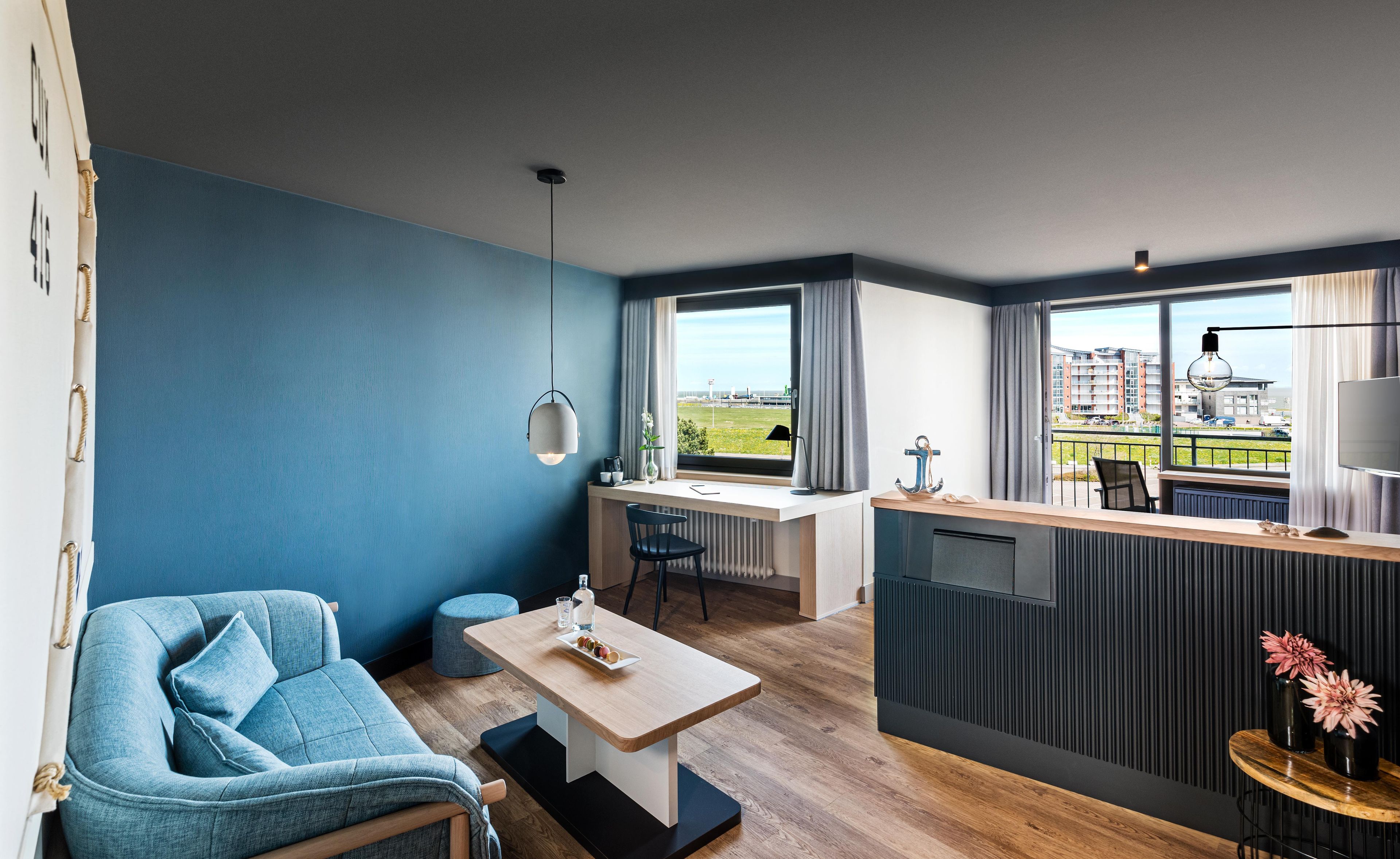Ein modernes Zimmer mit blauer Wand, einer Couch, einem Esstisch, einem Schreibtisch und Blick auf Gebäude durch große Fenster.