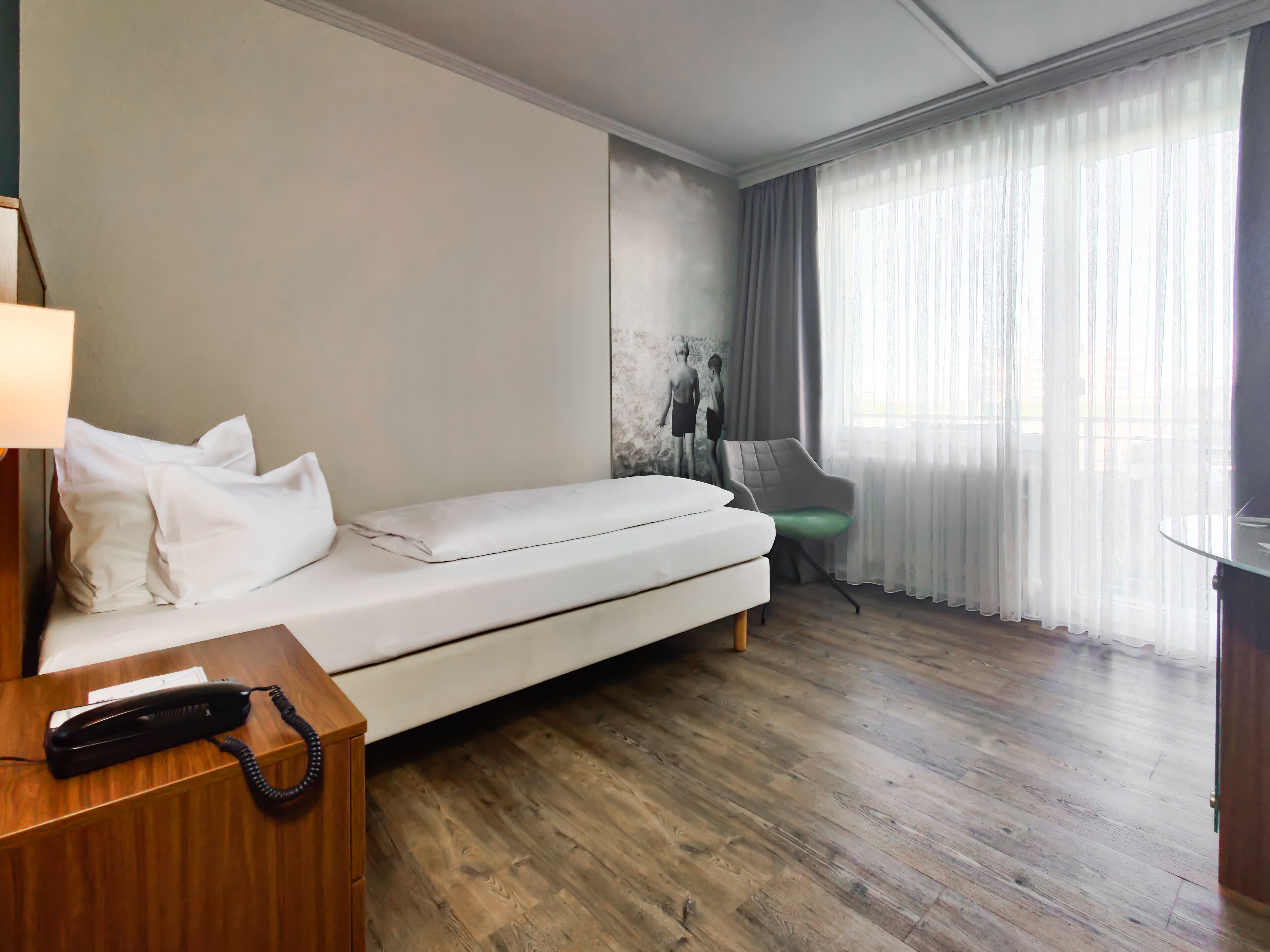 A hotel room with a single bed, a wooden nightstand with a telephone, a green chair, and a large window with sheer curtains.