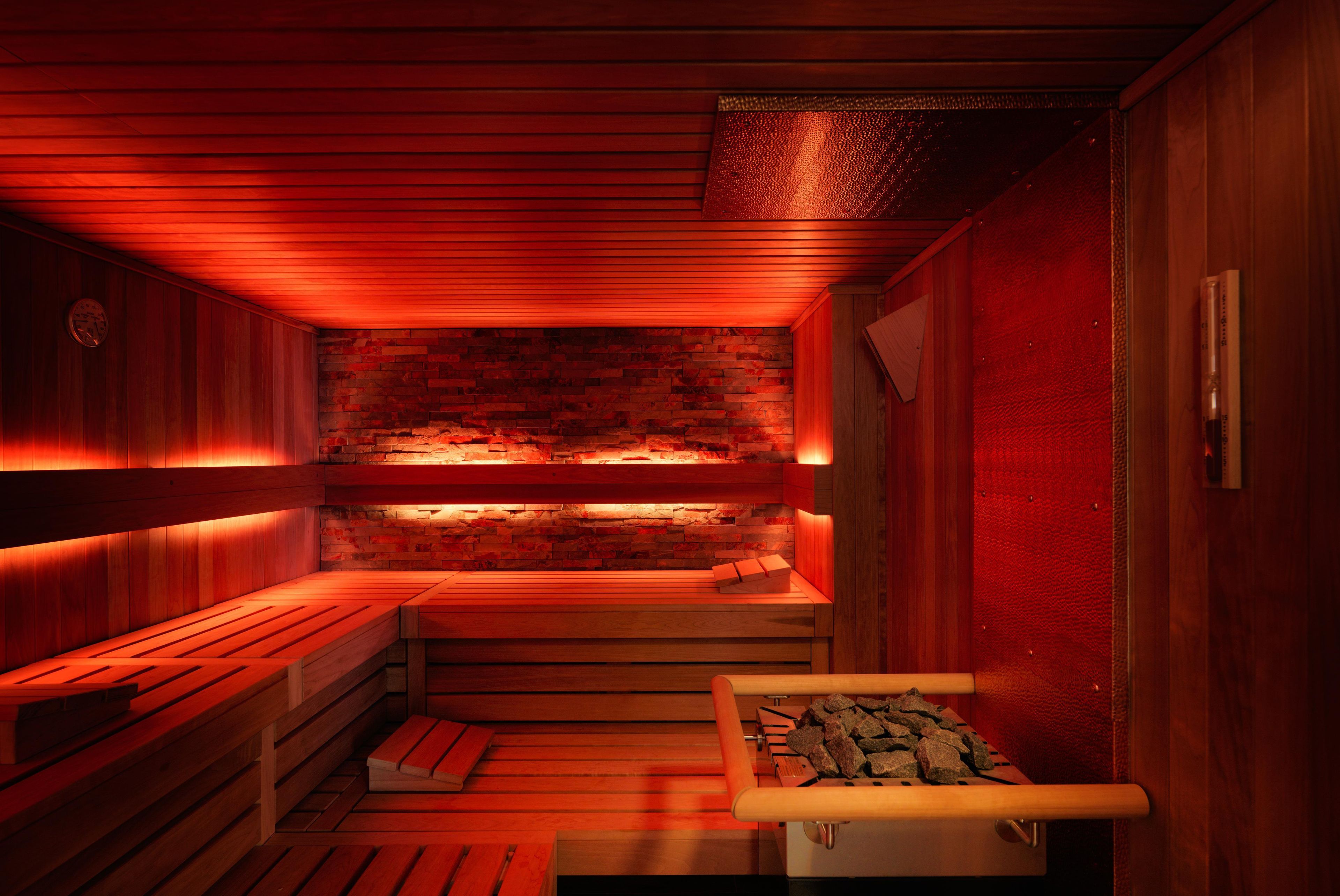 A sauna with wooden benches and a stone wall, equipped with a heater and a rock-filled container.