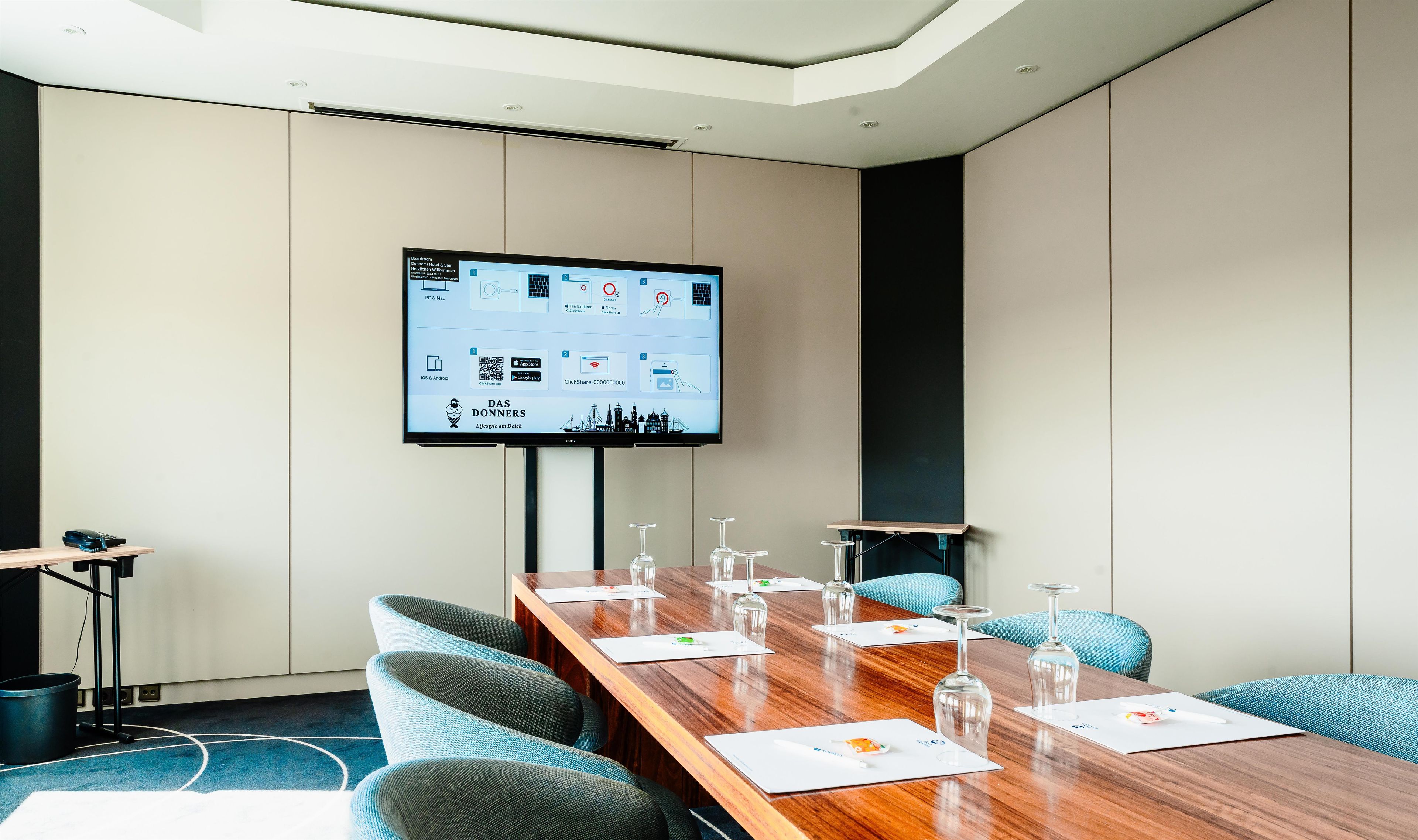 Ein Konferenzraum mit einem langen Holztisch, blauen Stühlen, einer Projektorleinwand und einer Whiteboard-Tafel.