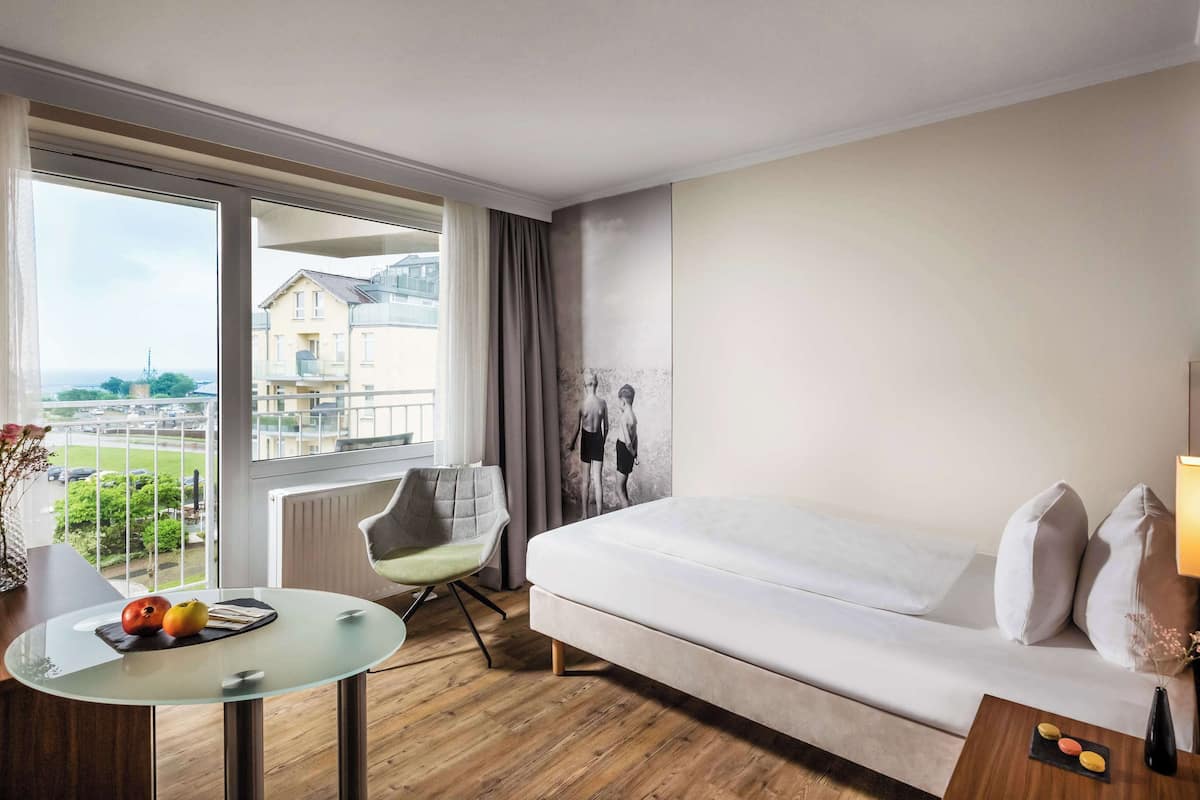 A hotel room with a bed, a small table with a vase, a chair, and a view of buildings and greenery outside.