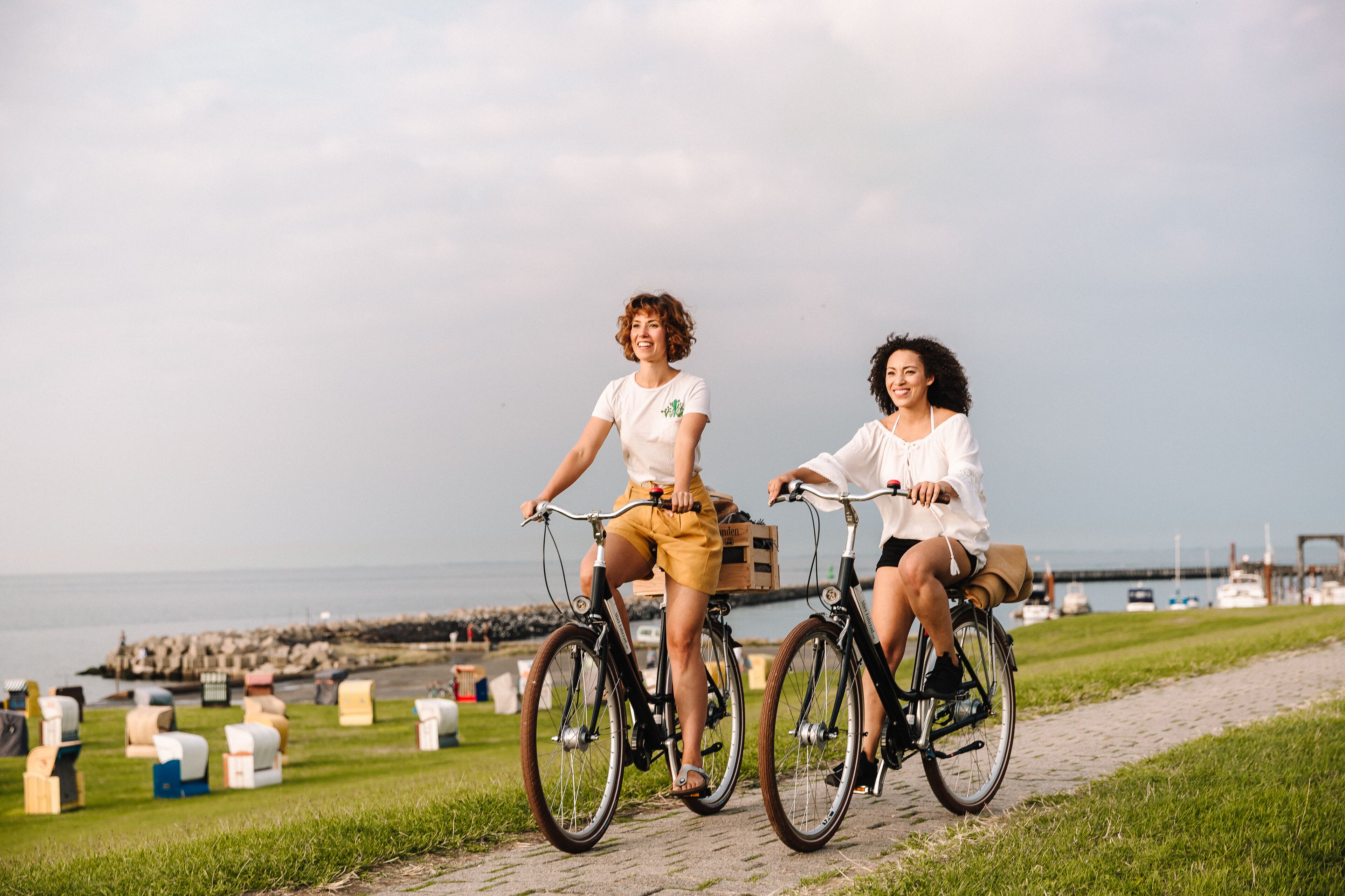 Two people riding bicycles on a paved path with a grassy area and a body of water in the background.