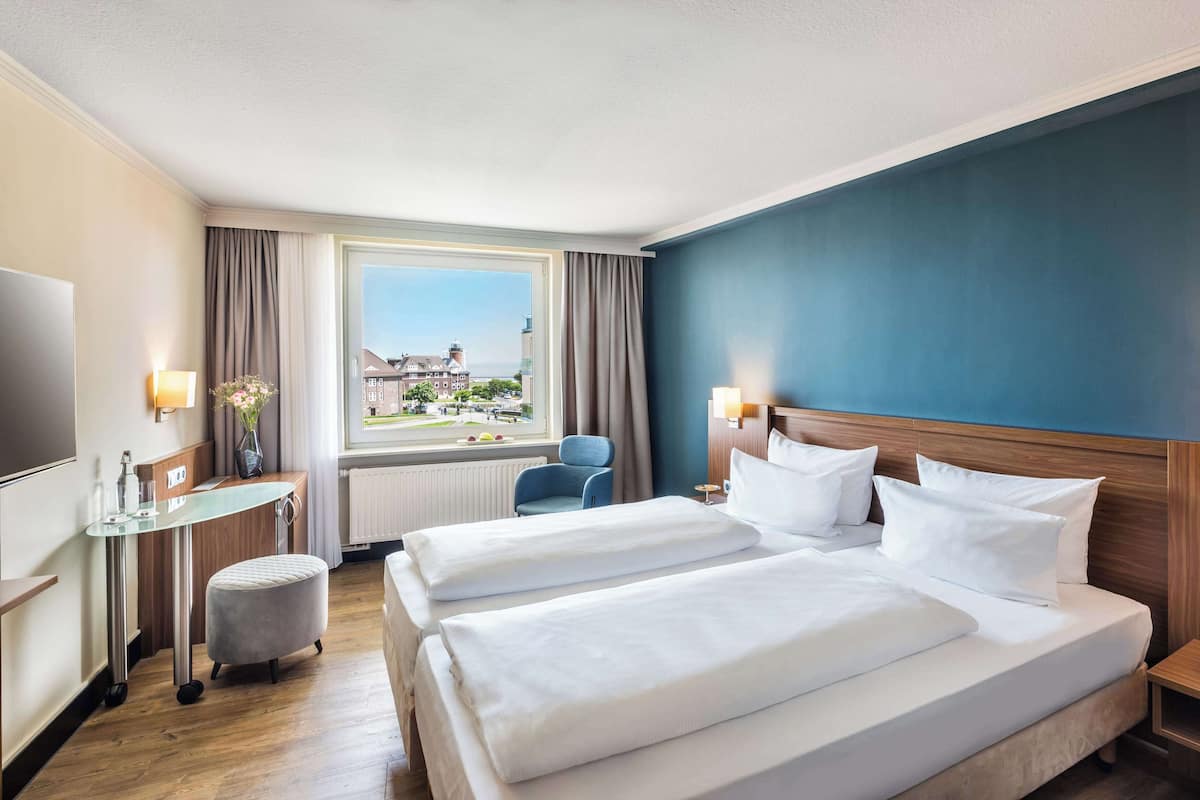 A hotel room with two beds, a desk, a chair, a TV, and a window with a view of buildings and a body of water.