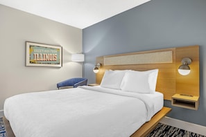 A hotel room with a large bed, a headboard with built-in lighting, a bedside table, a wall-mounted lamp, and a framed sign on the wall.