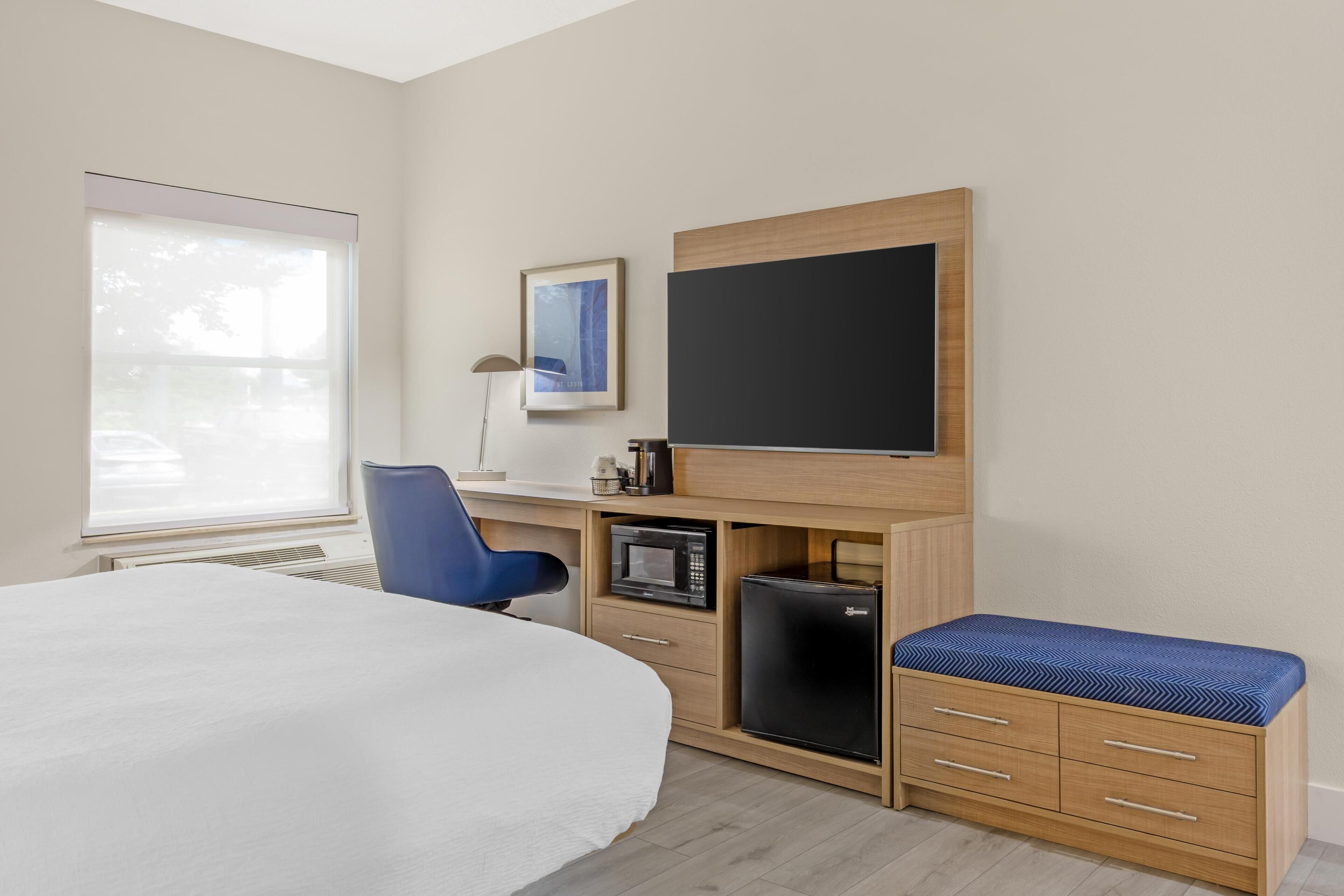 A hotel room with a bed, a desk with a computer, a microwave, a refrigerator, a chair, and a large window.
