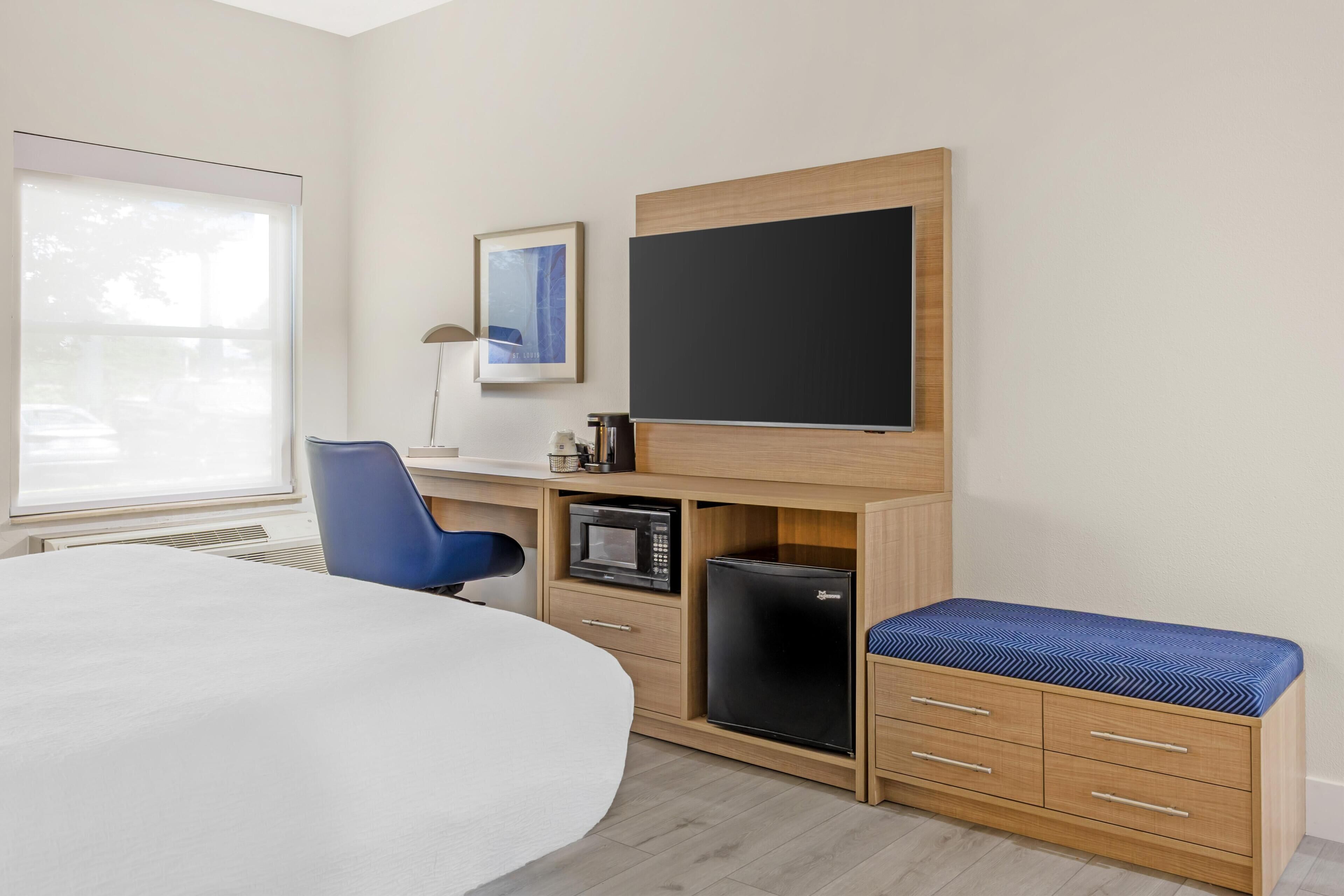 A hotel room with a bed, a desk with a computer, a small refrigerator, a television, and a window.