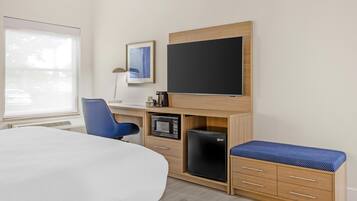 A hotel room with a bed, a desk with a computer, a small refrigerator, a television, and a window.