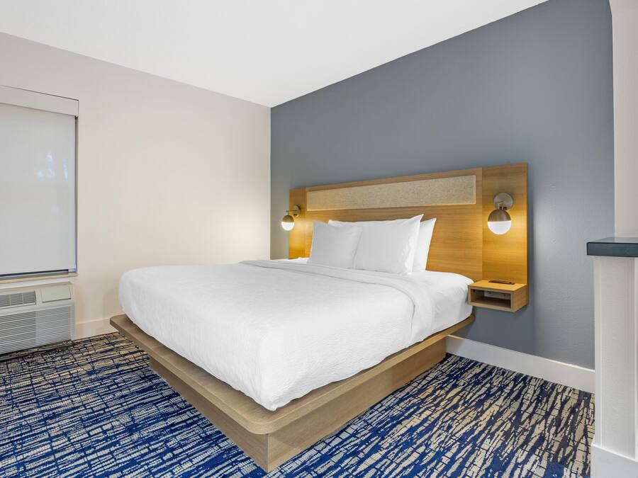 A hotel room with a bed, a headboard with wall-mounted lights, a white wall, and a blue patterned carpet.