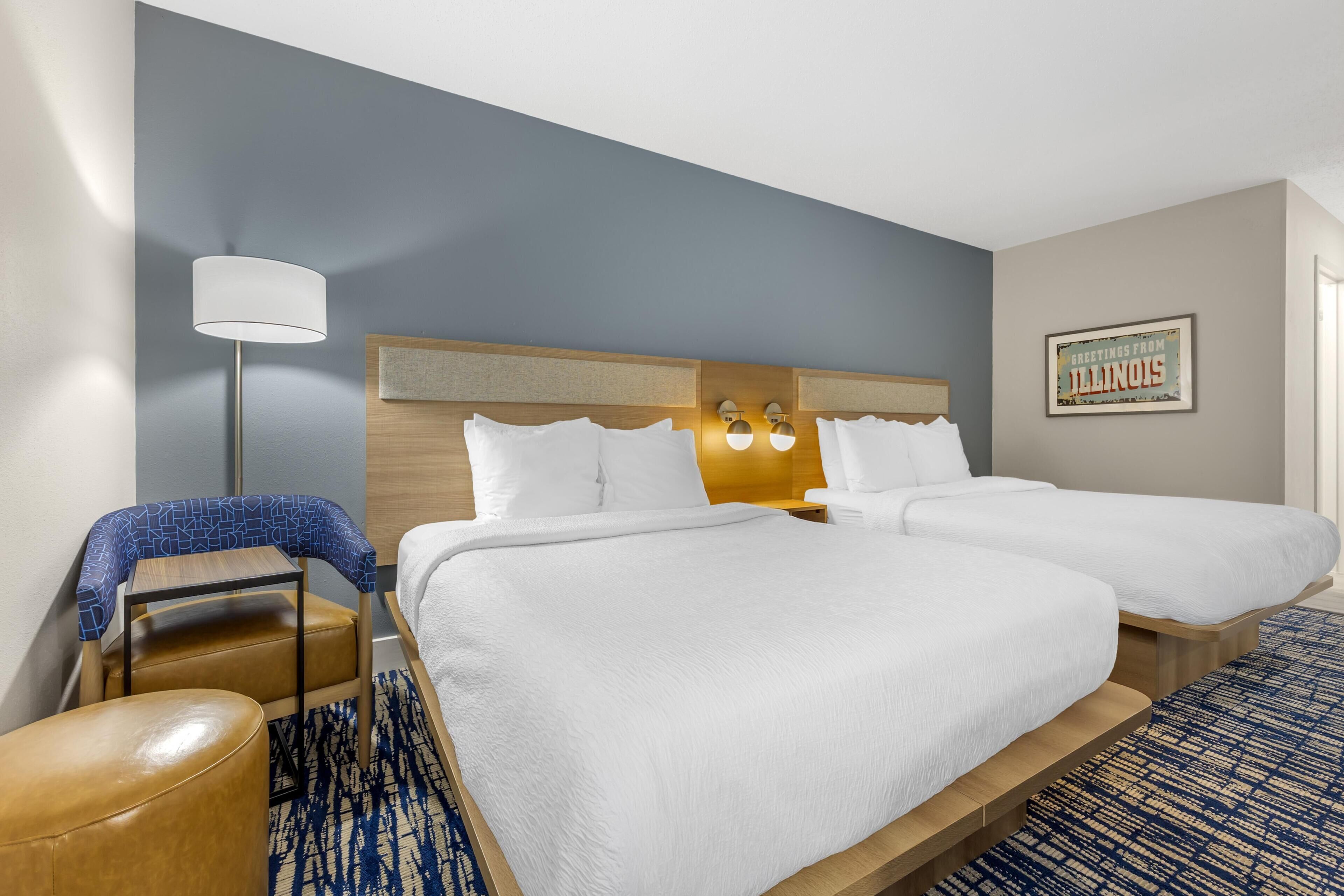 A hotel room with two beds, a desk, a chair, a lamp, and a wall-mounted light fixture.