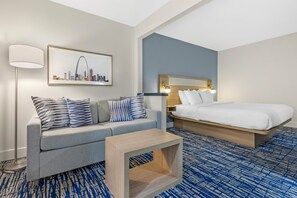 A hotel room with a bed, a sofa, a coffee table, a lamp, and a cityscape painting on the wall.