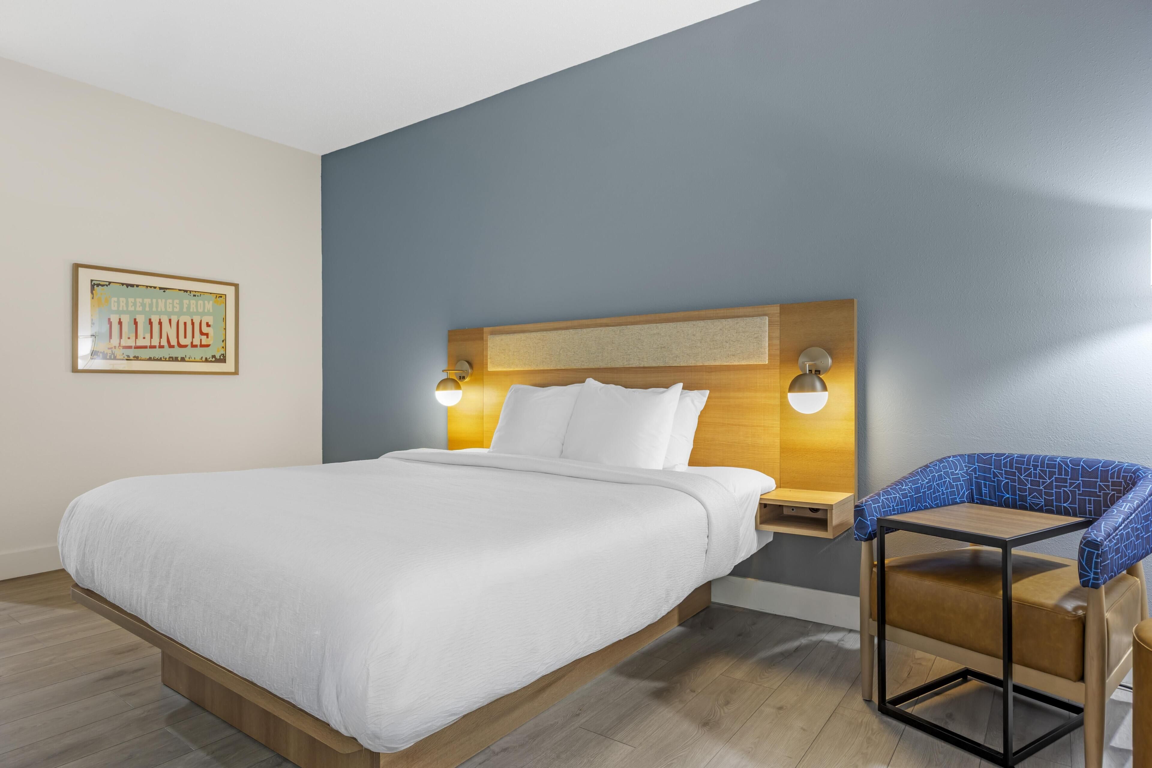 A hotel room with a bed, bedside tables, a chair, and a wall-mounted light fixture.