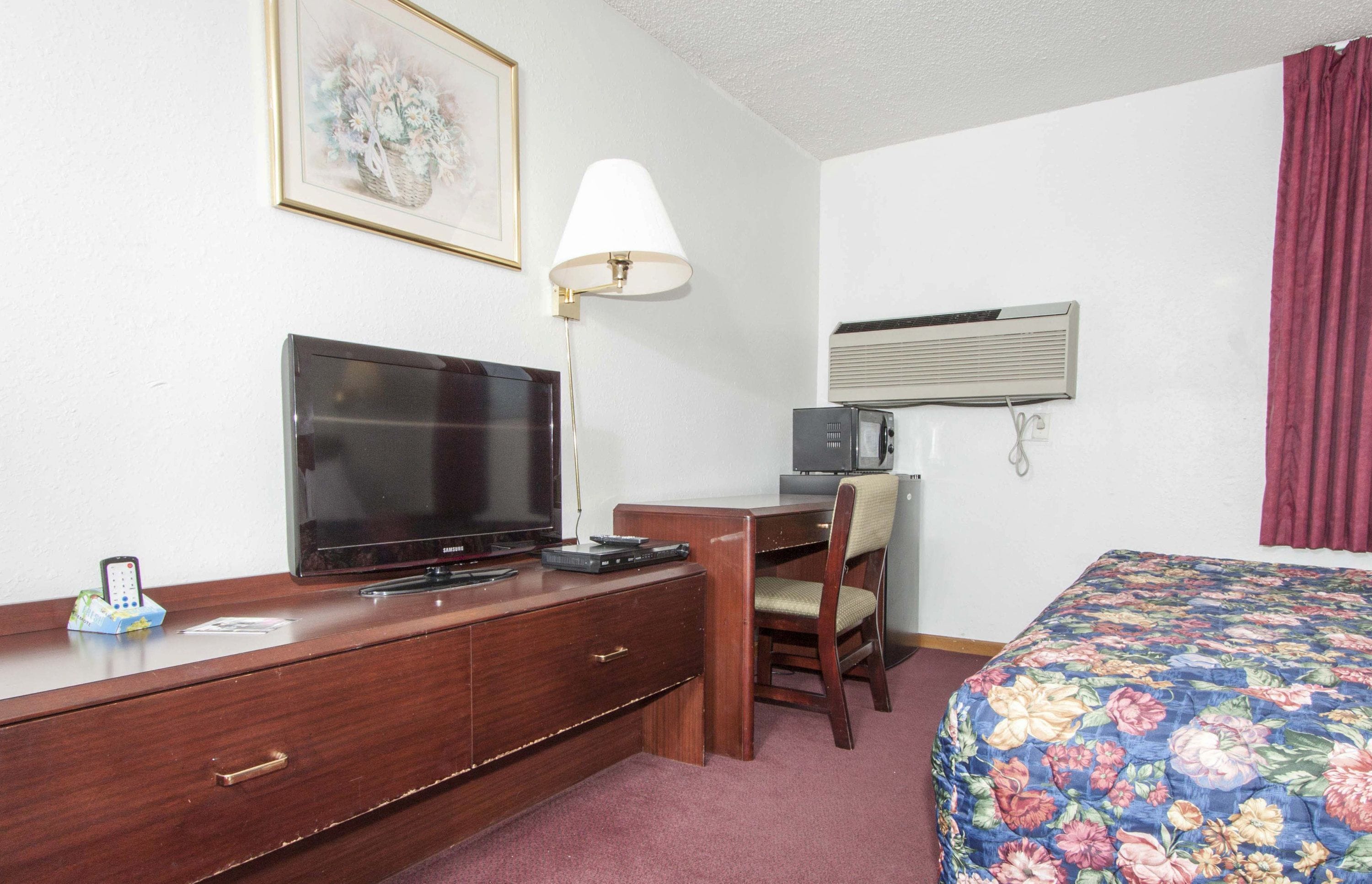A hotel room with a bed, a desk with a television, a lamp, and an air conditioning unit.