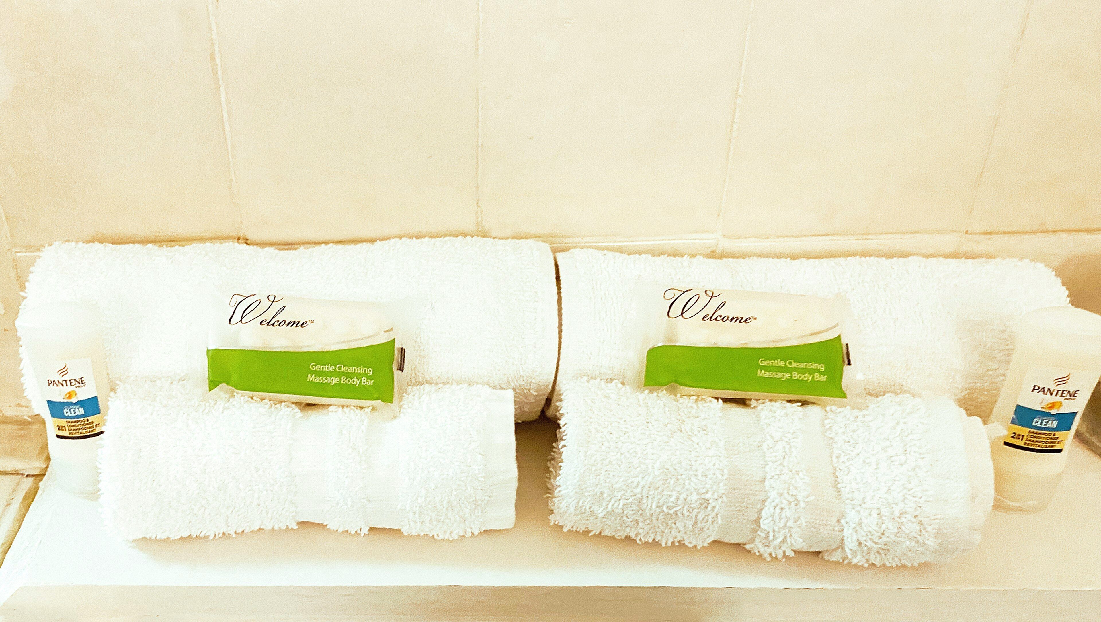 Two neatly folded towels with a green bar in the center and a white label on the left, placed on a white surface against a beige wall.