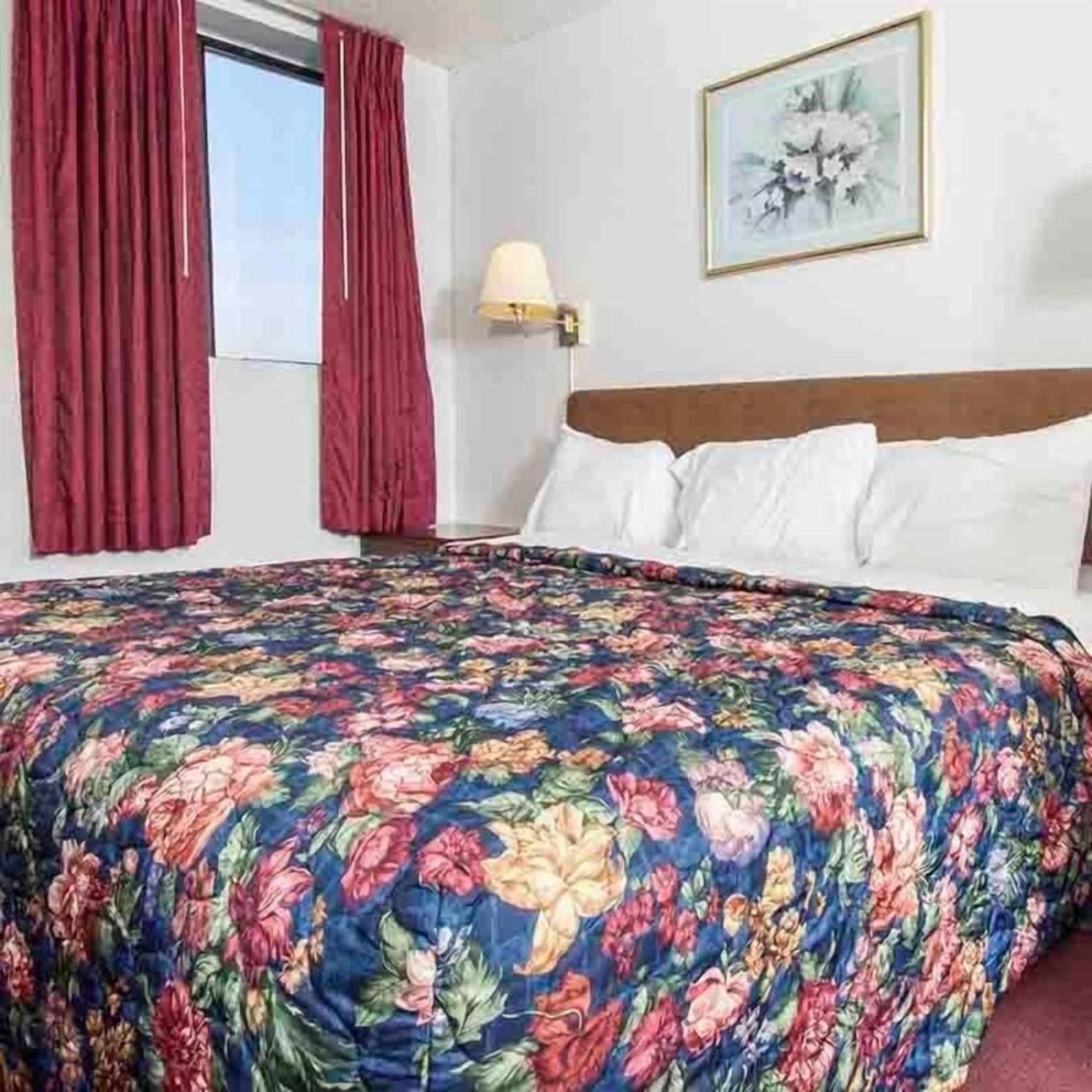 Hotellihuone, jossa on kukkakuviollinen päiväpeite, puinen pääty, valkoiset tyynyt, punaisilla verhoilla varustettu ikkuna ja seinään kiinnitetty lamppu.