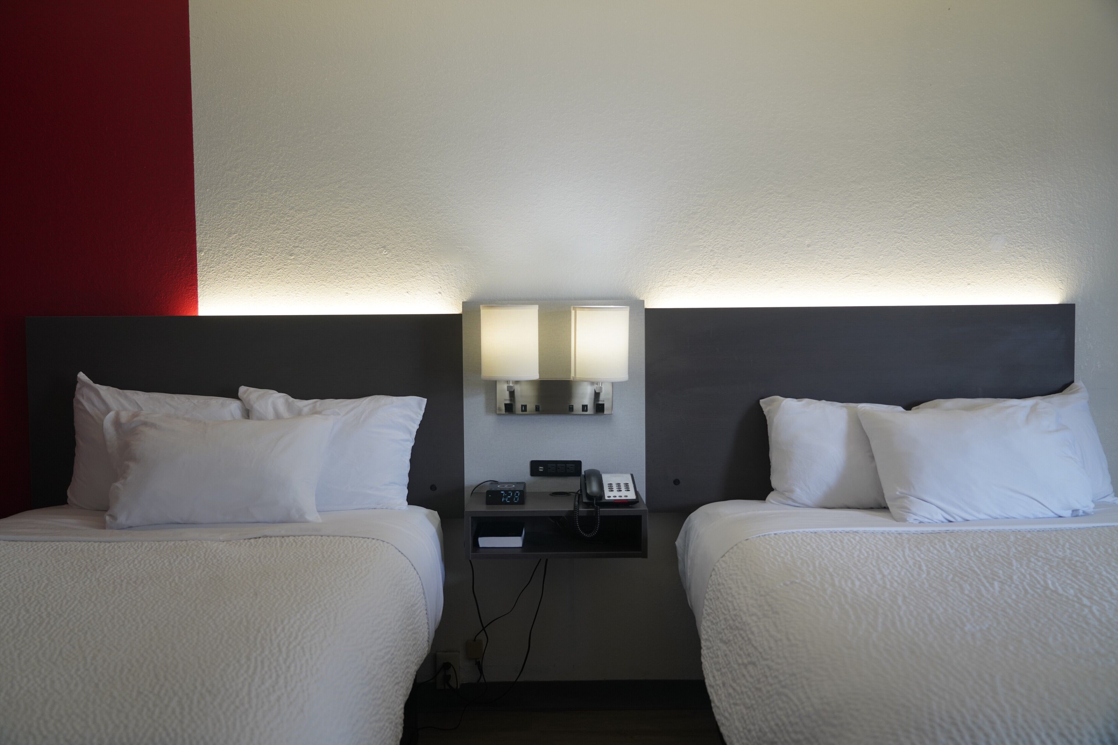 A hotel room with two beds, a nightstand with a phone and clock, and a wall-mounted light fixture.