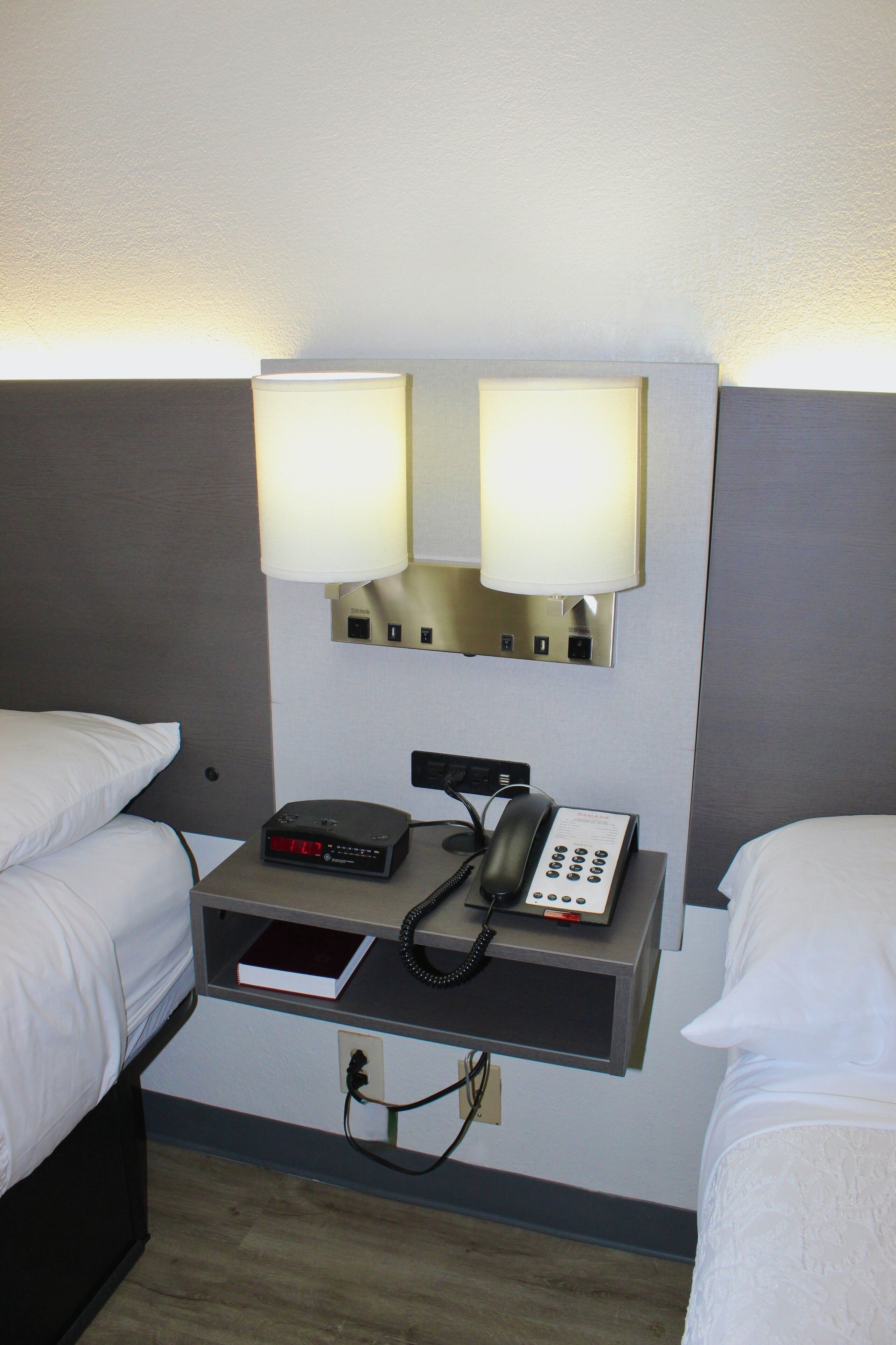 A hotel room with two beds, a bedside table with a phone and a digital clock, and a wall-mounted lamp.