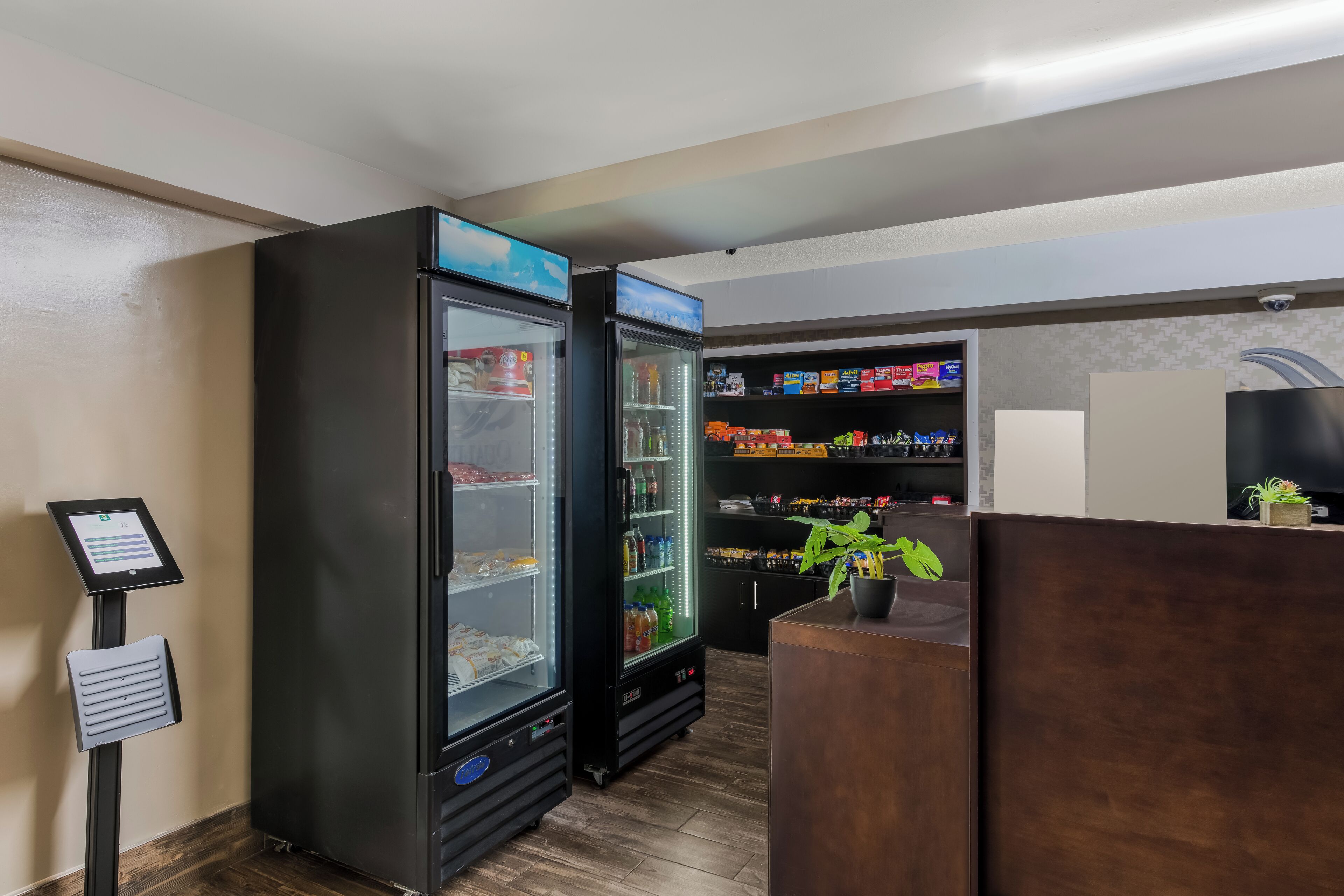 A hotel lobby with two large black refrigerators, a digital kiosk, and a reception desk with a potted plant.
